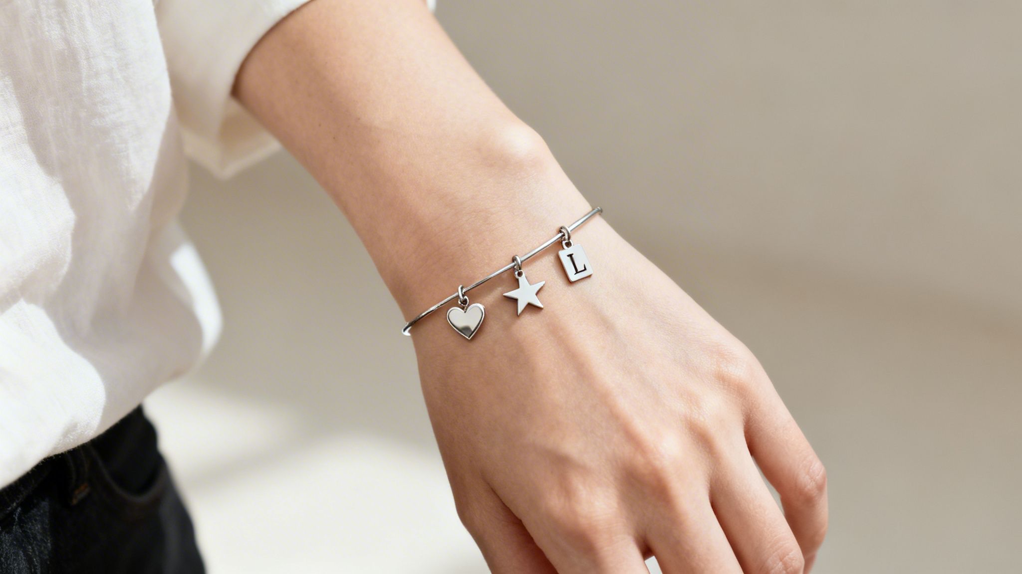 A close-up of a silver charm bracelet on a woman's wrist, featuring heart, star, and 'L' letter charms.