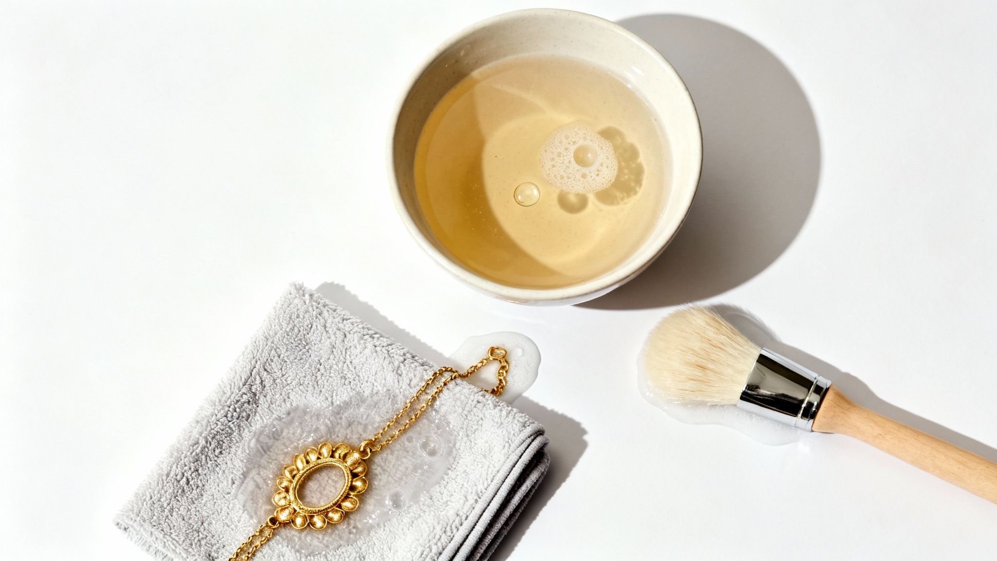 Gold necklace, cleaning solution, brush, and towel laid out for jewelry care on a white surface.