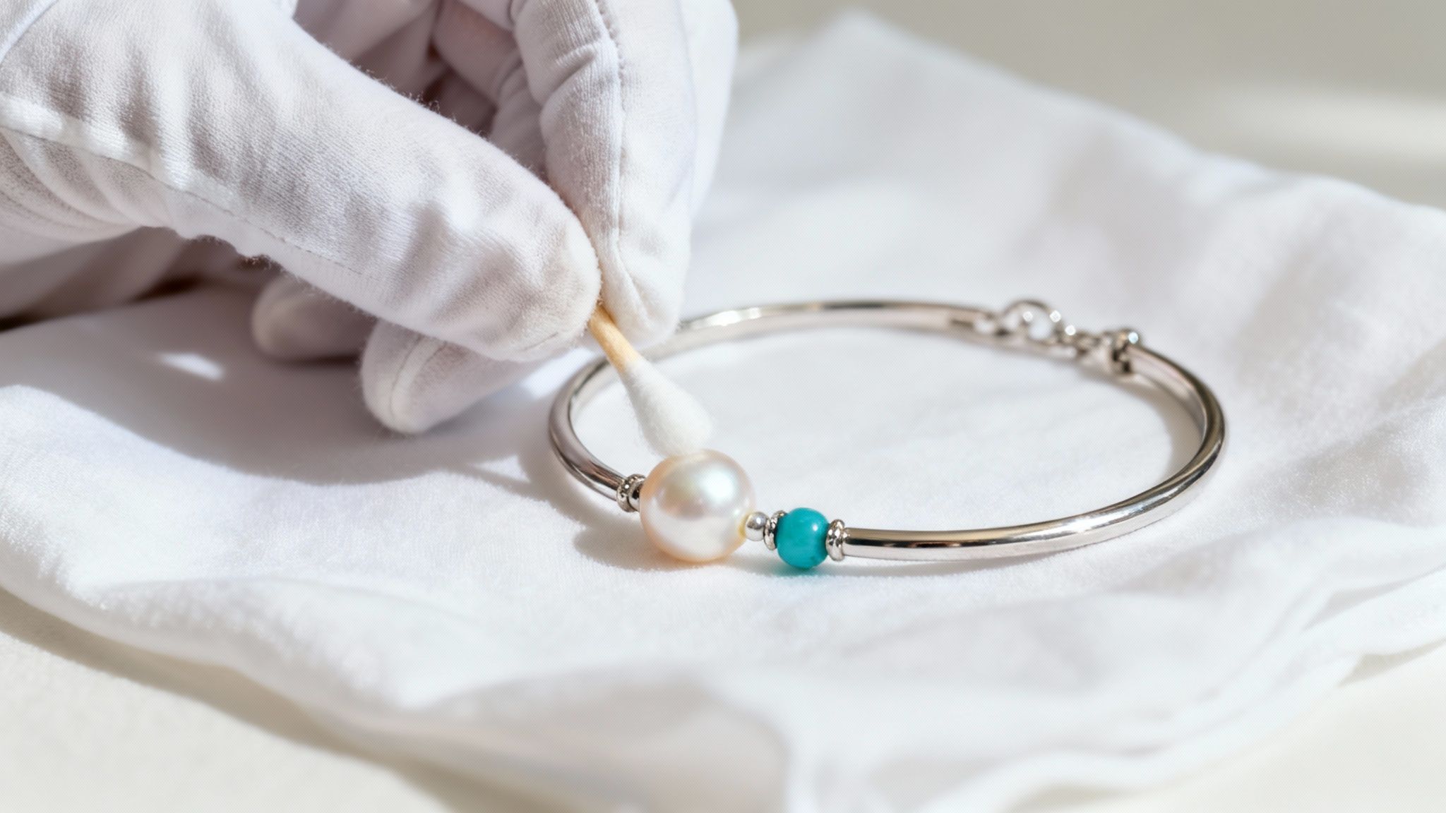 A gloved hand gently cleans a silver bracelet with a pearl and turquoise bead using a cotton swab on a white cloth.