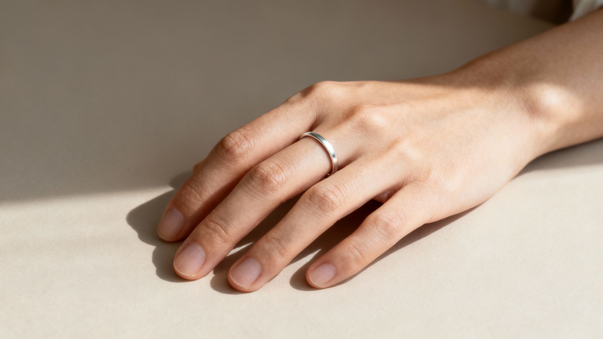 A hand with a simple silver ring on the ring finger, resting on a beige surface with shadows.