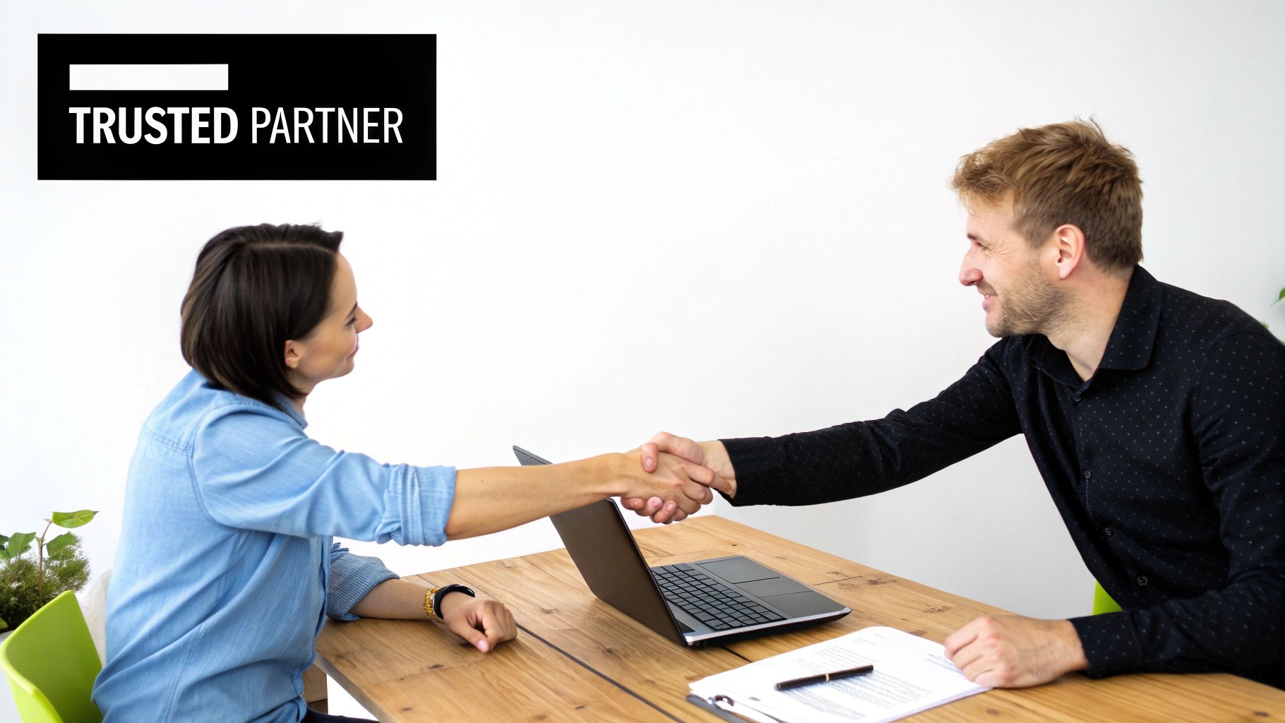 Two business partners shake hands across a table with a laptop, signifying trust and collaboration.
