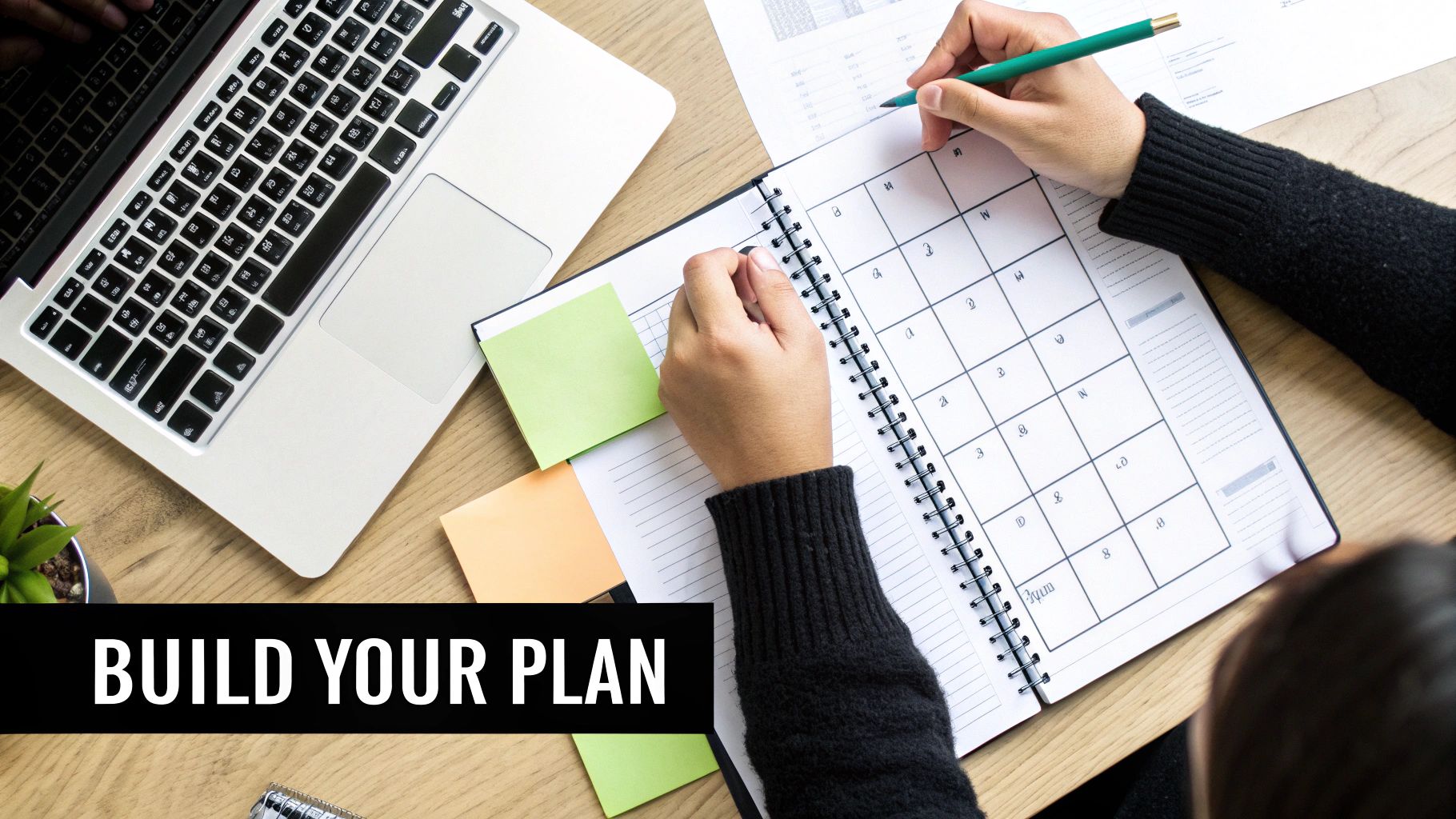 Overhead view of a person writing in a planner with a pencil, laptop, and sticky notes, emphasizing planning.