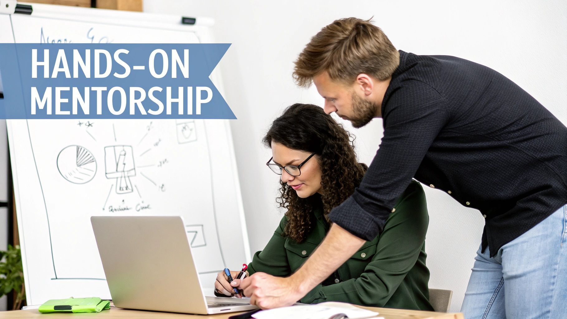A man provides hands-on mentorship to a woman working on a laptop with a whiteboard.