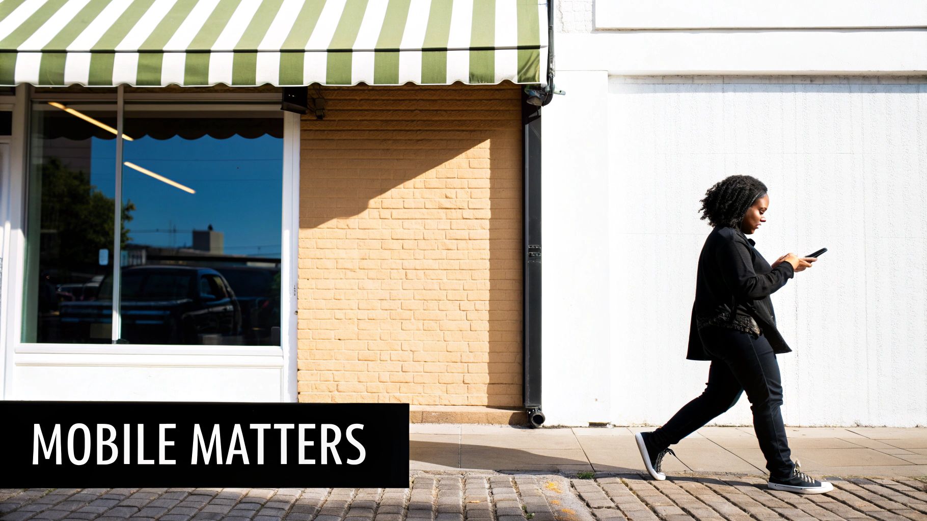 A young Black woman walks on a city sidewalk, looking down at her smartphone.