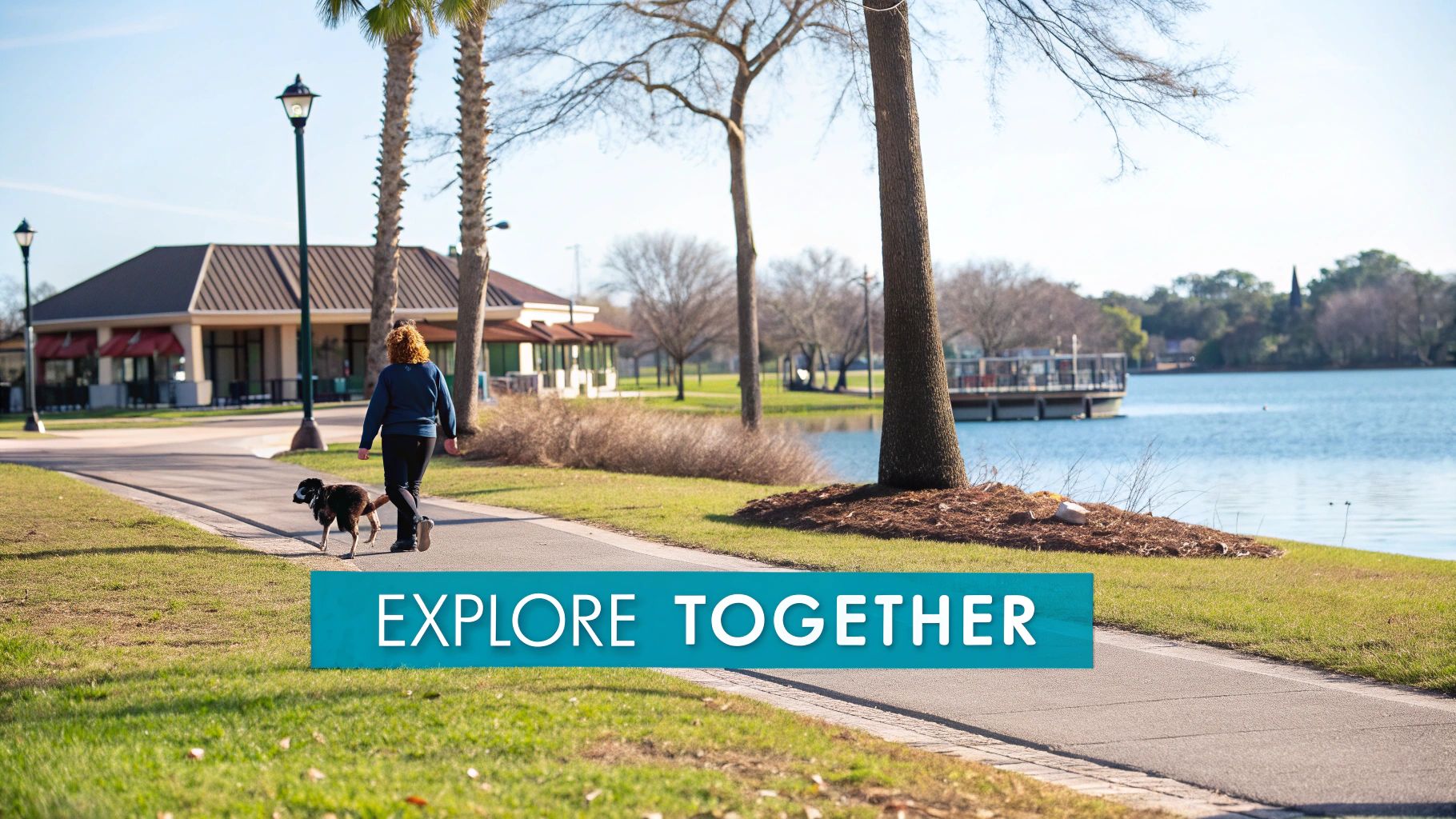 A person walks their dog on a paved path next to a lake with buildings and trees.