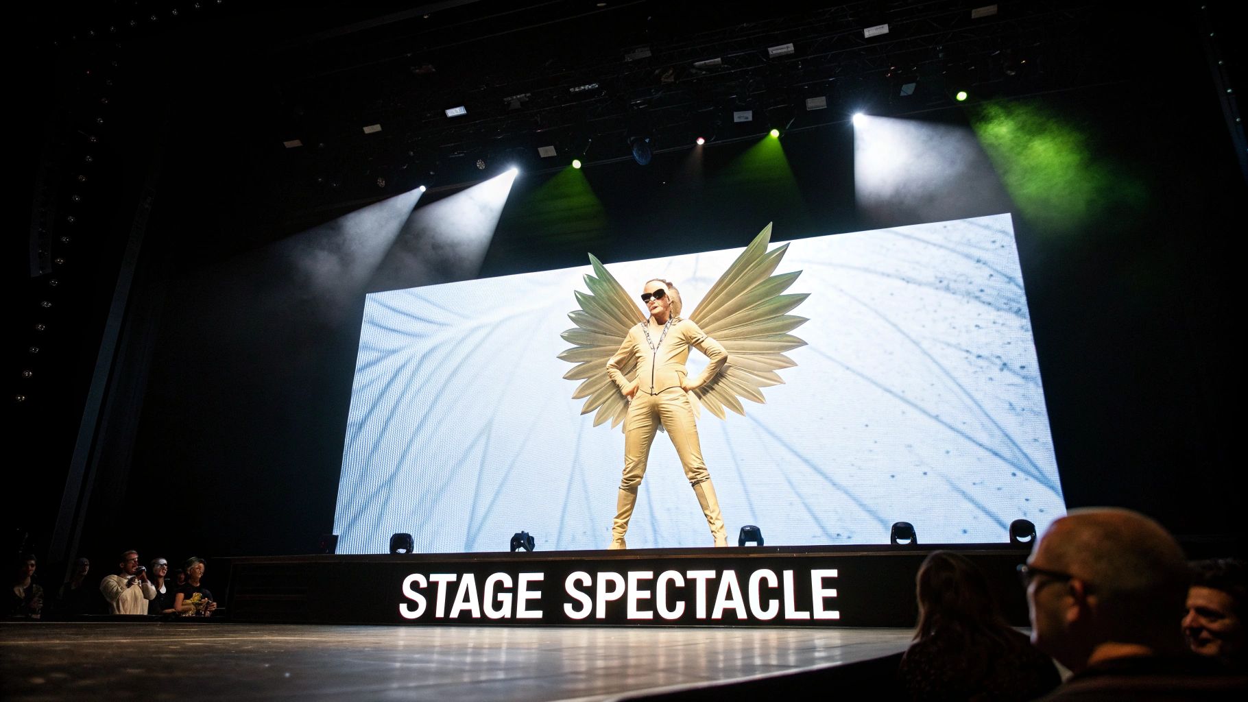 A performer in a striking gold winged outfit and sunglasses stands on a stage during a concert.