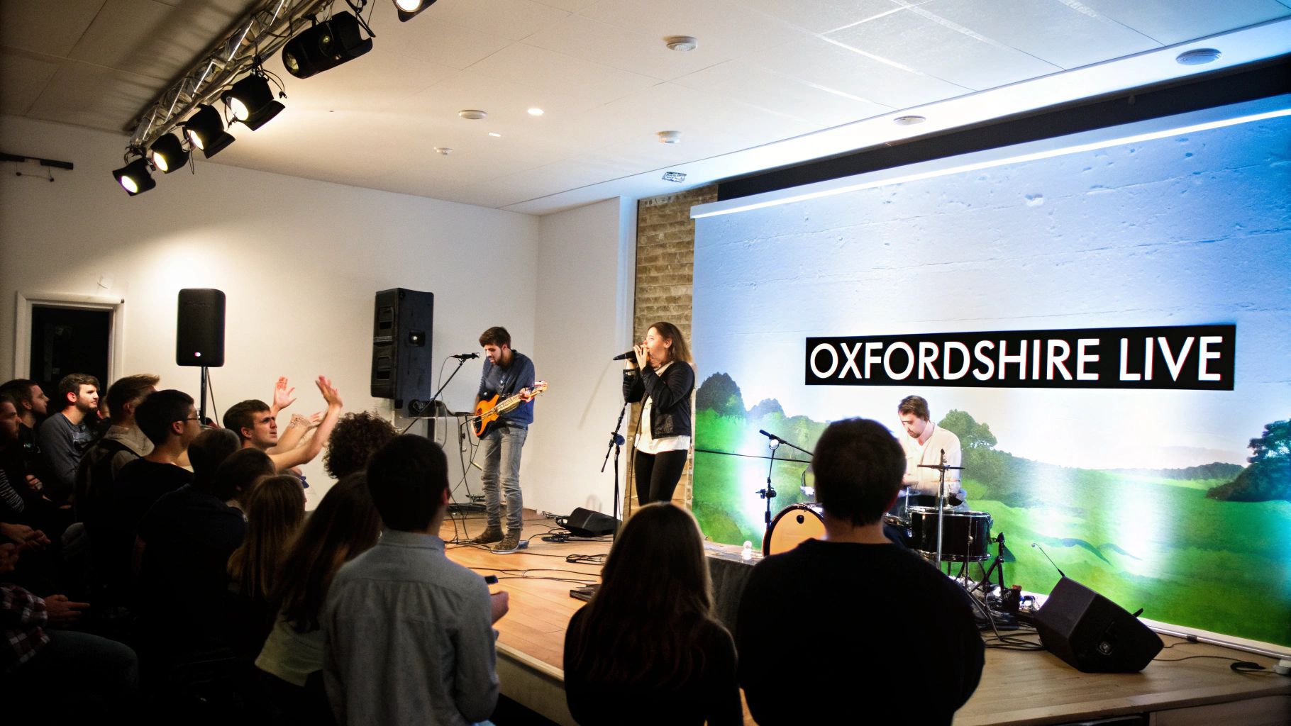 A band performs live music on stage, featuring a singer, guitarist, and drummer, for an engaged audience with a 'OXFORDSHIRE LIVE' screen.