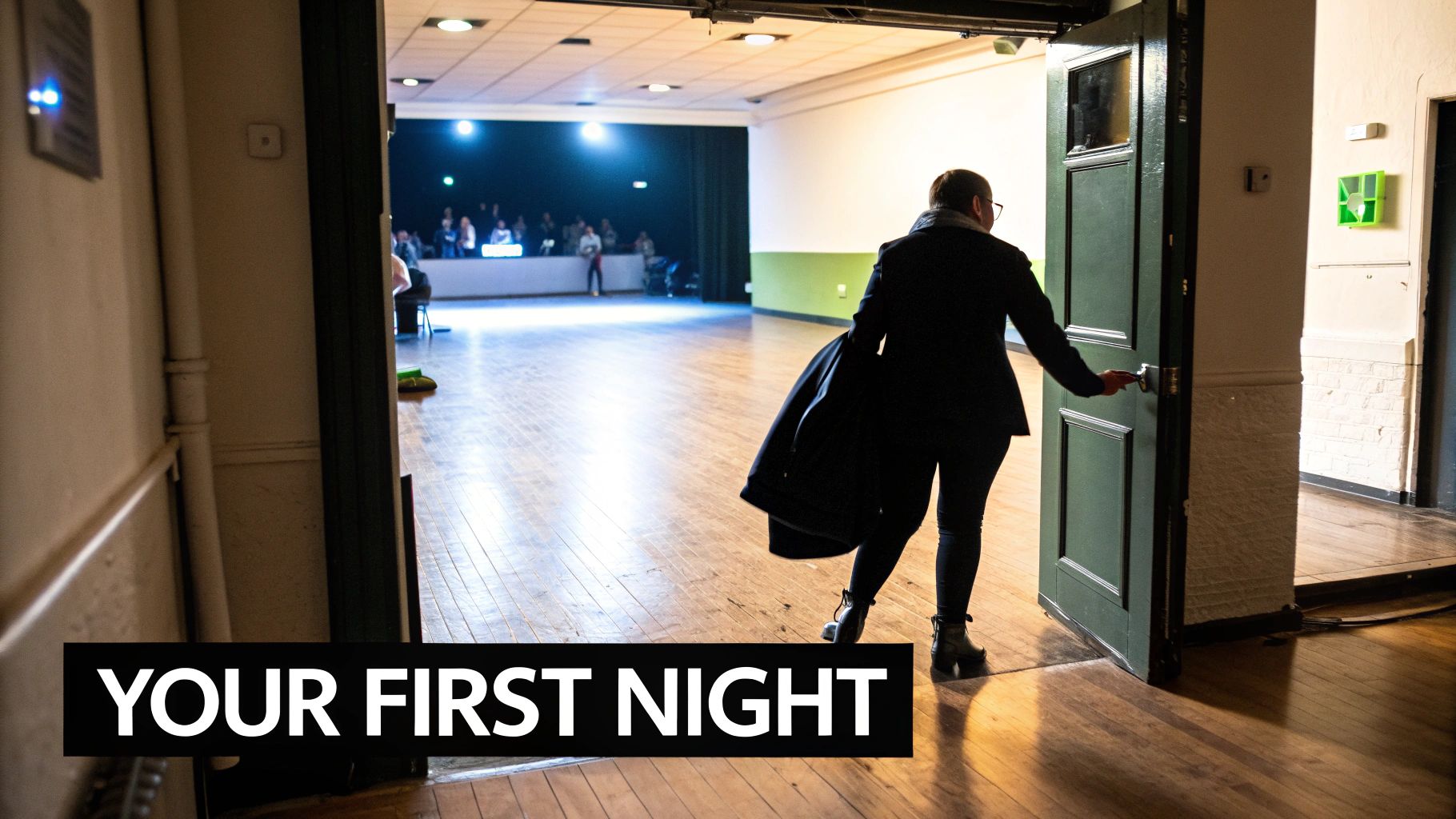 A person in a coat opens a dark green door, entering a brightly lit hall with a wooden floor and a distant stage.