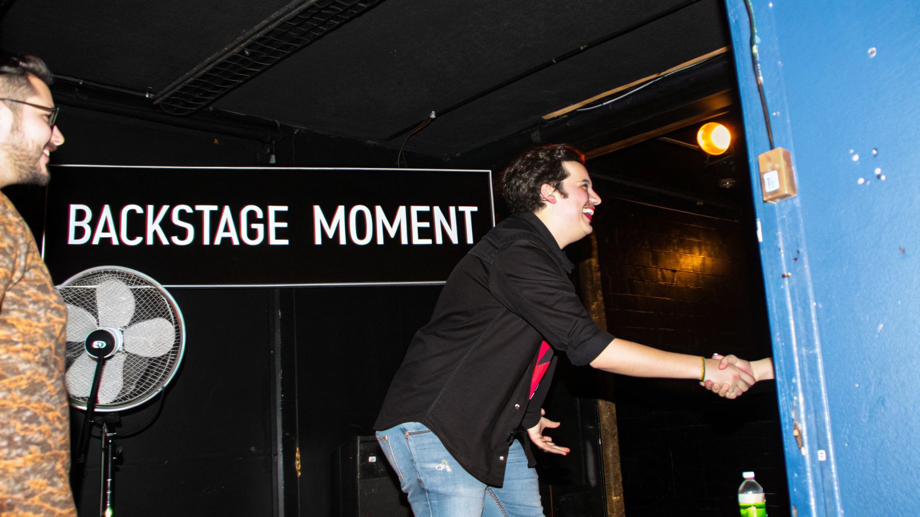A man in a black shirt smiling and shaking hands with someone off-camera, next to a 'BACKSTAGE MOMENT' sign.