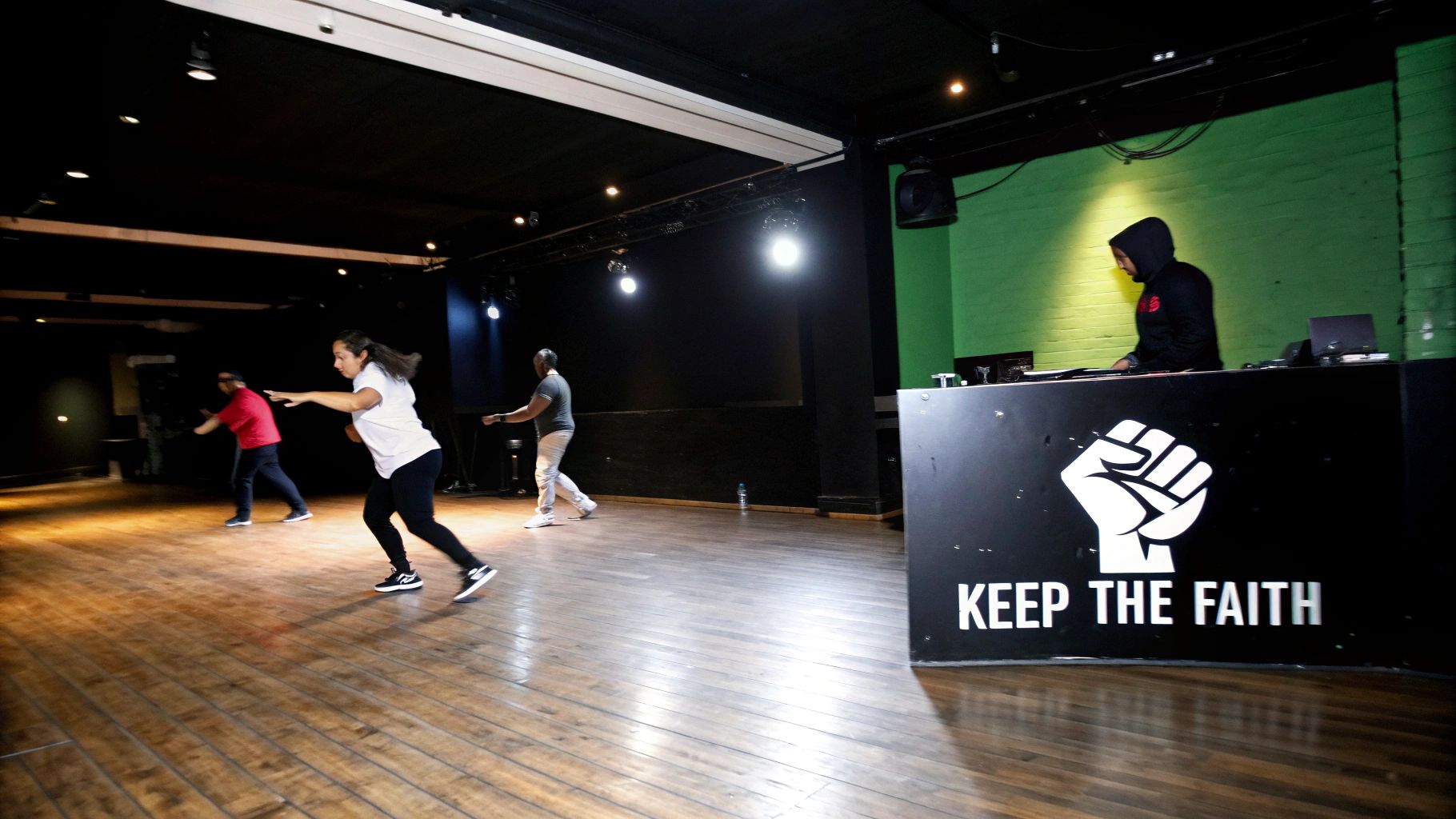 People dancing in a spacious room with a wooden floor and a DJ booth featuring 'KEEP THE FAITH' text.