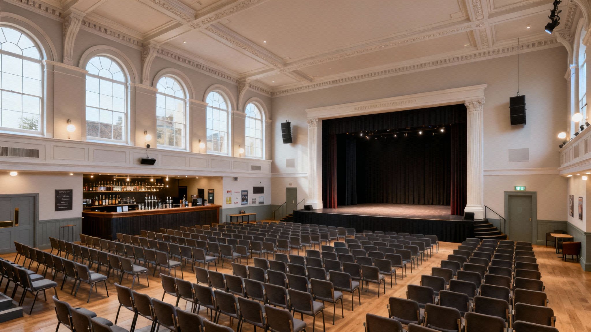 Spacious event hall with a stage, black curtains, rows of chairs, and a bar area.
