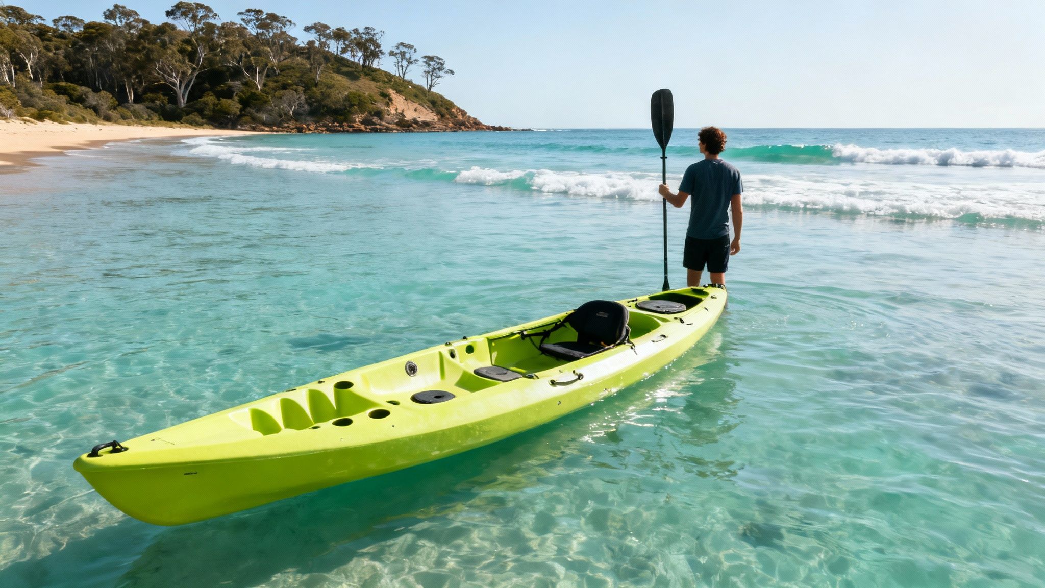 A person standing in clear turquoise water beside a green sit-on-top kayak, looking at the waves.