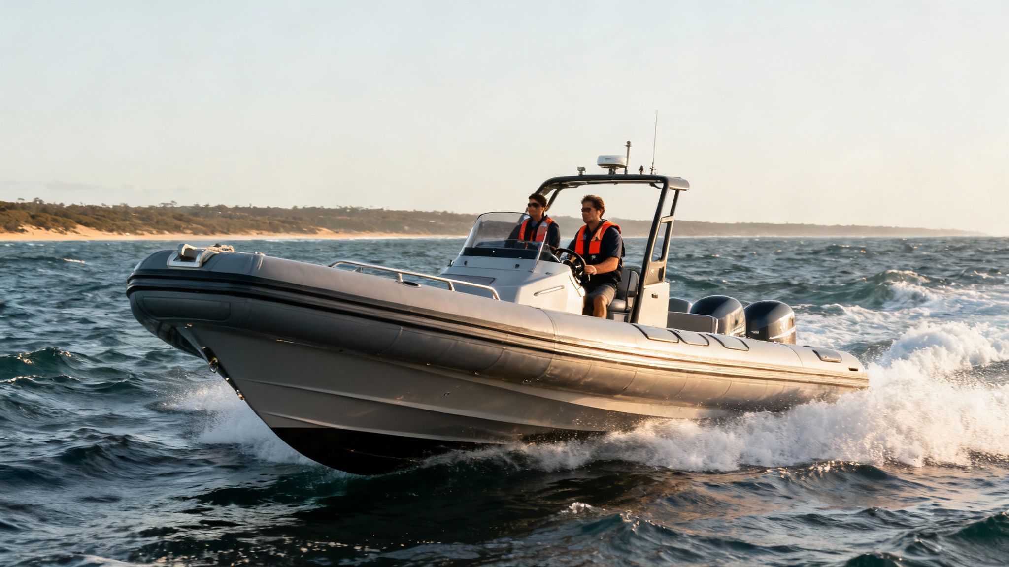 Two men in life jackets drive a grey rigid inflatable boat fast on choppy ocean waters near a sandy beach.