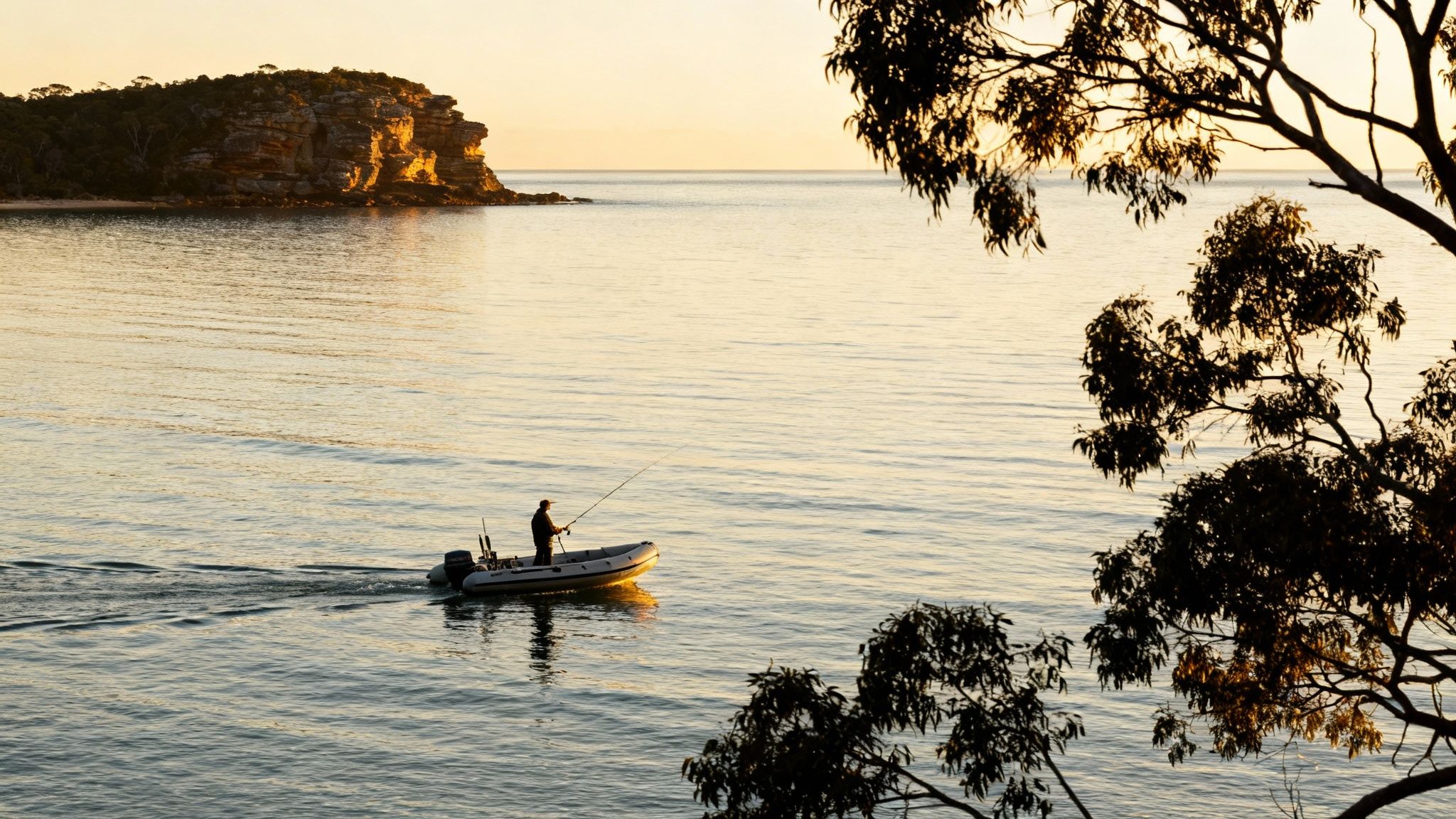 A person fishes from an inflatable boat on serene water at sunset, with a rocky island and trees.