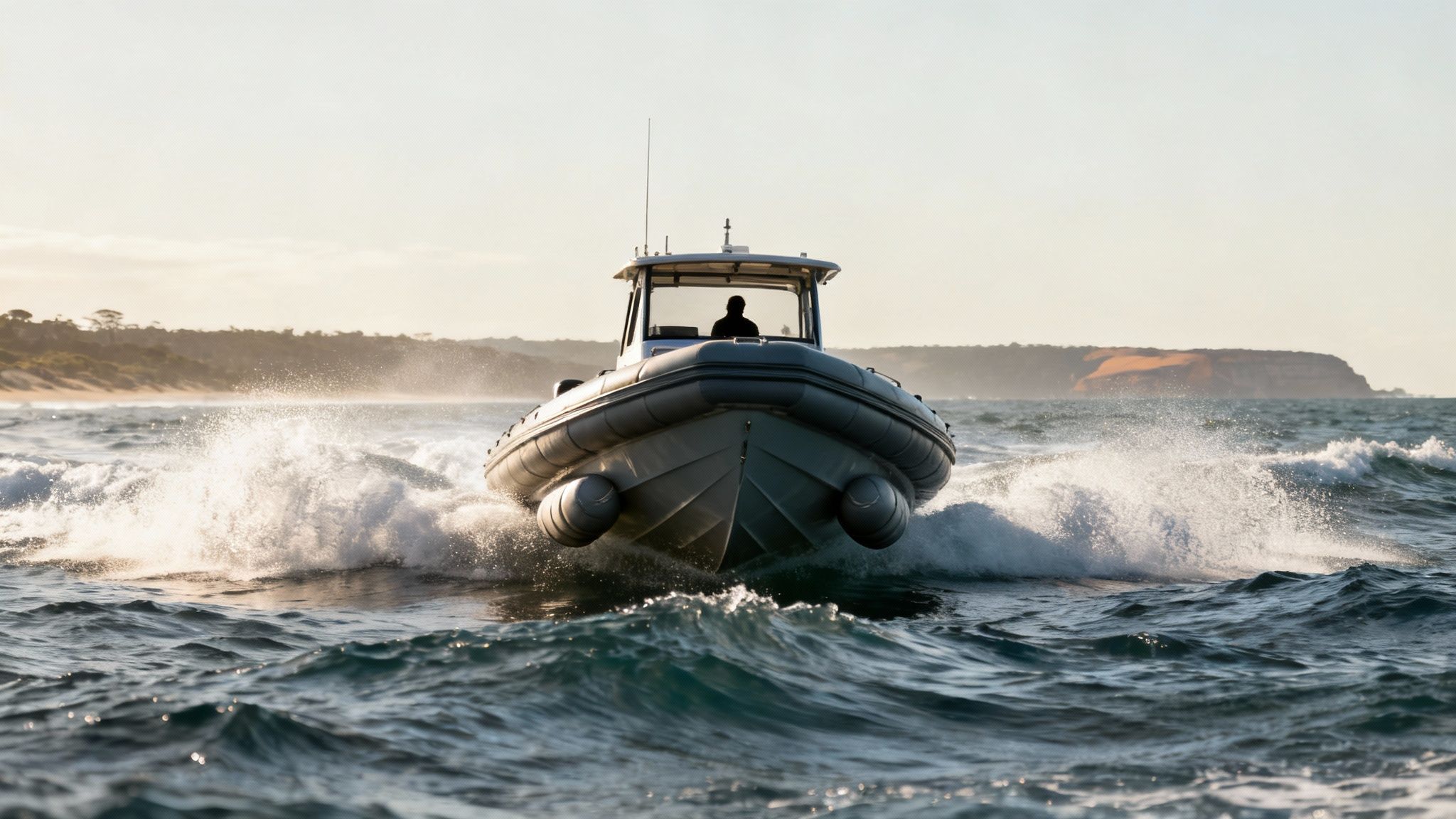 A grey rigid hull inflatable boat speeds through choppy ocean water, creating white splashes, with a coastline in the background.