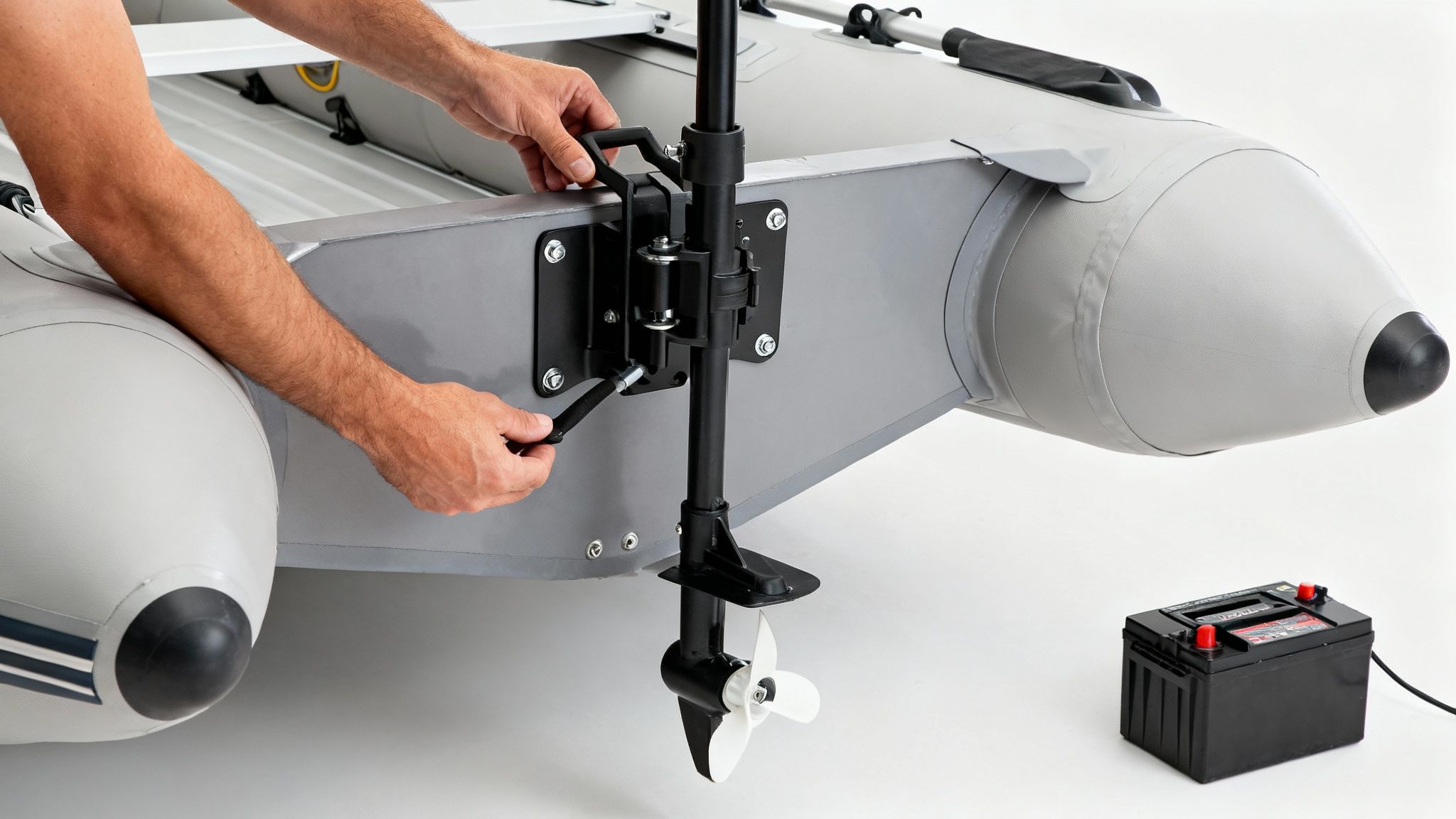 Hands are installing an electric trolling motor on the transom of an inflatable boat, with a battery nearby.
