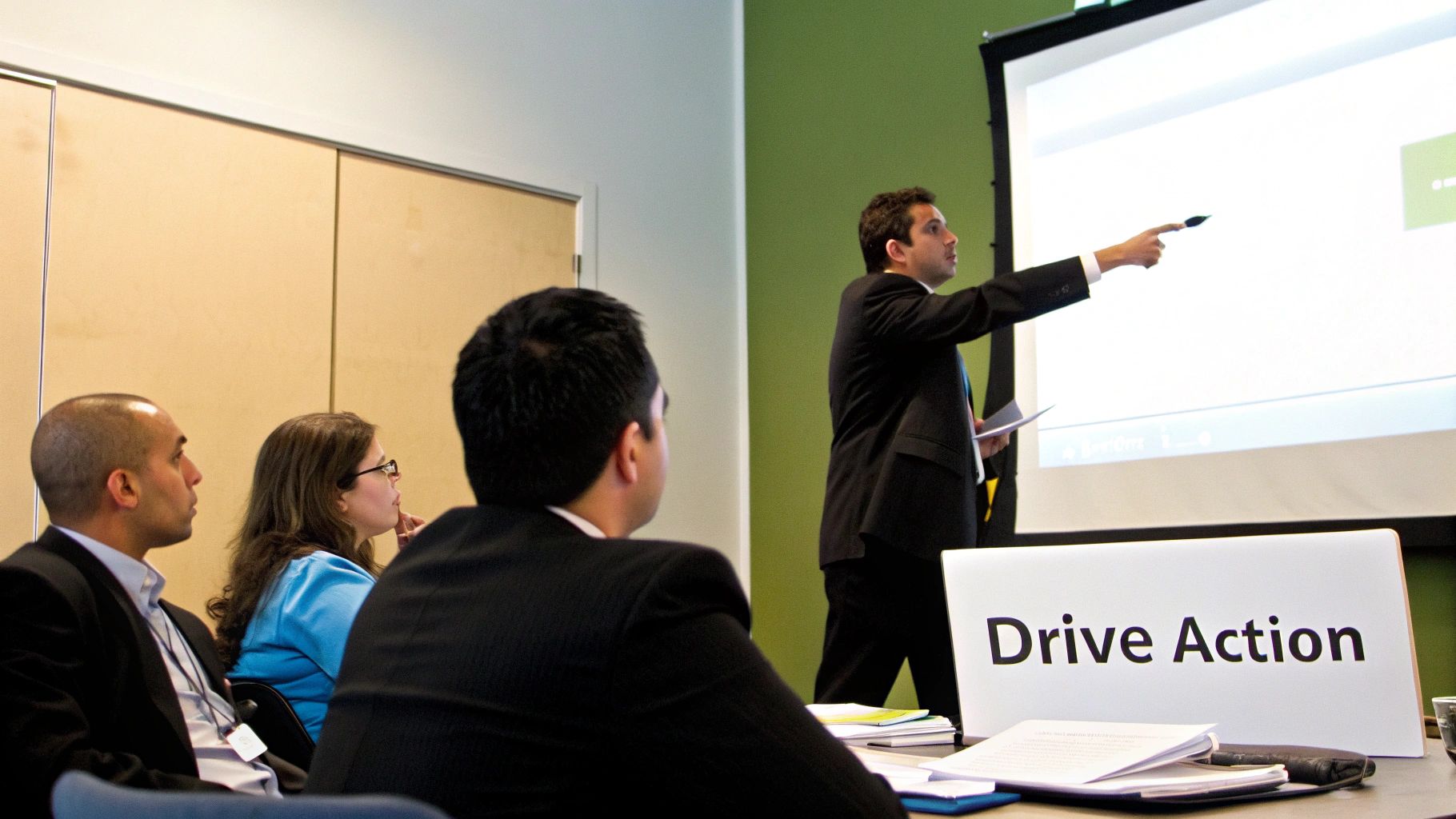 A speaker in a suit points at a presentation screen during a business meeting with seated attendees.