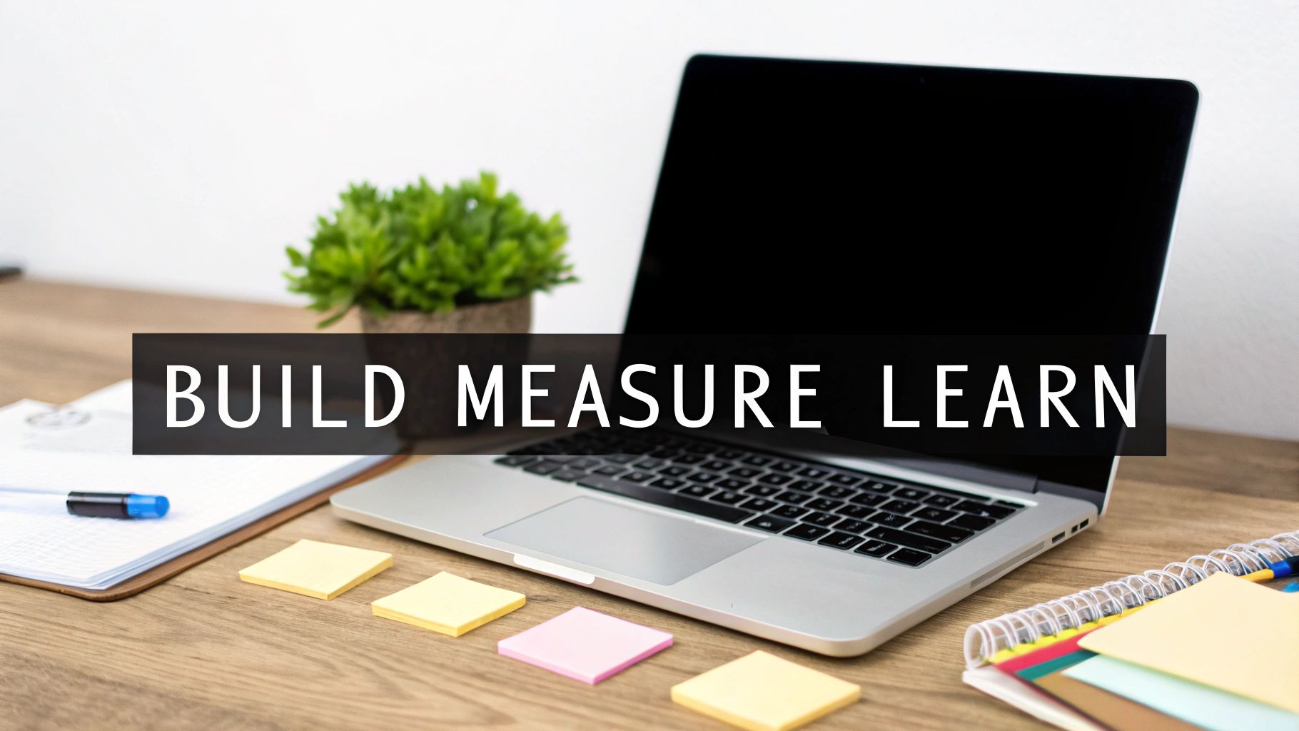 A modern workspace with a laptop, plant, and notepad, featuring a 'BUILD MEASURE LEARN' banner.