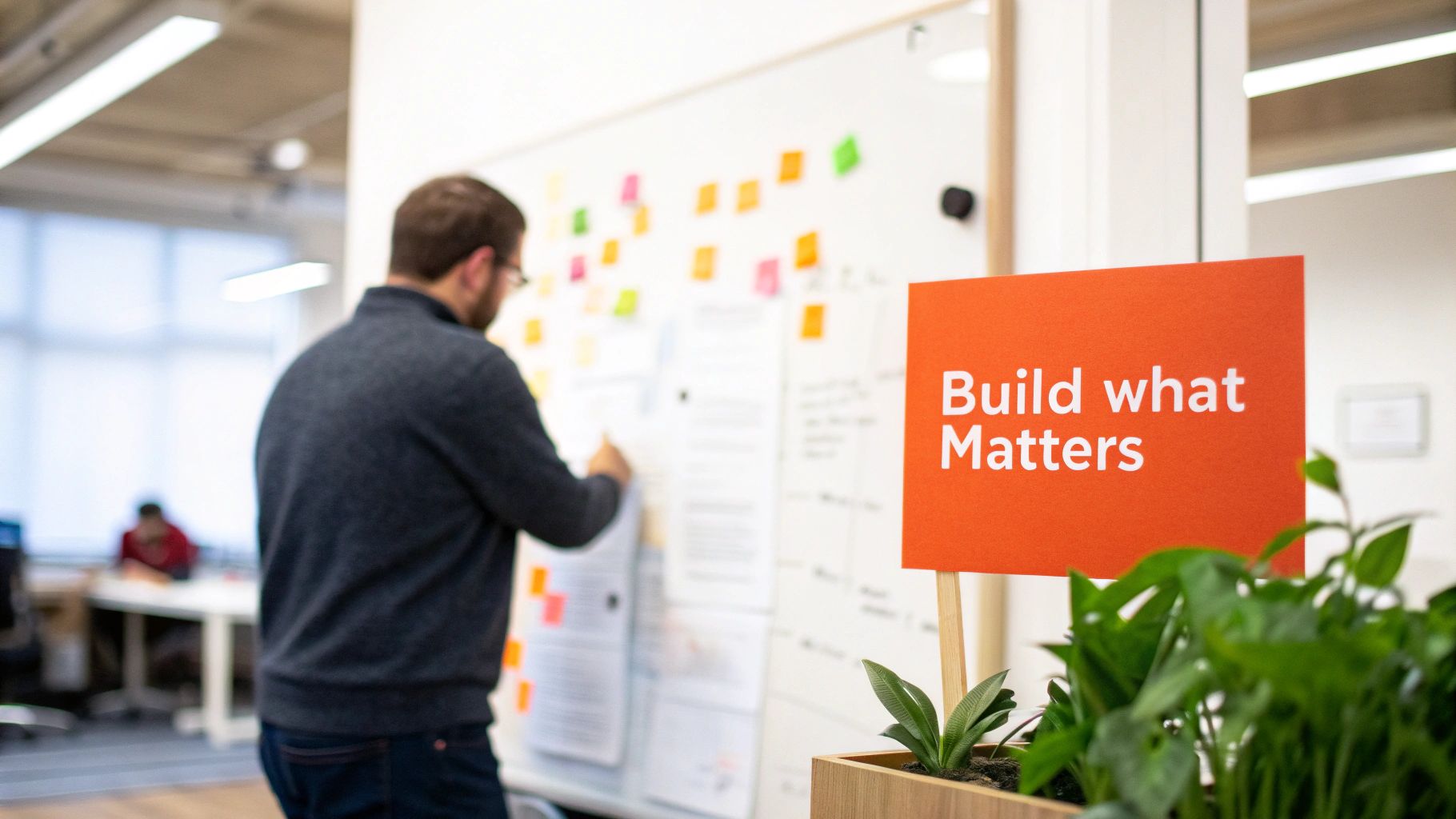 A man in an office looking at a whiteboard with sticky notes, next to a plant with a "Build what Matters" sign.