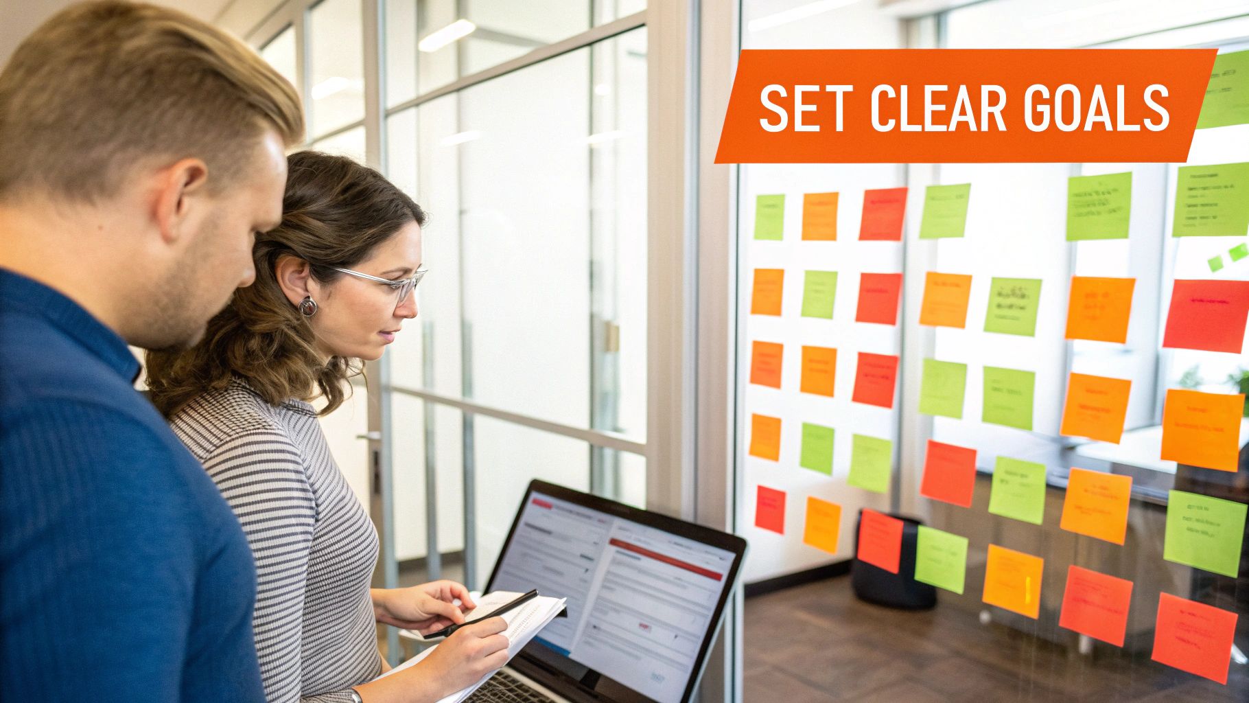 Two colleagues planning in an office, looking at a laptop with sticky notes on a glass wall.