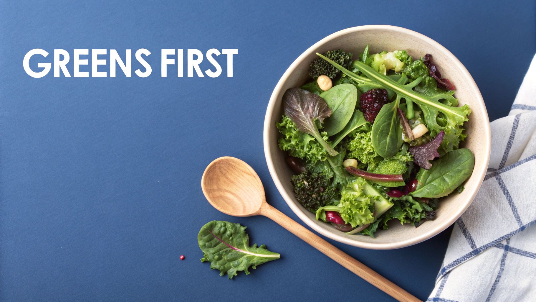 Overhead shot of a fresh green salad in a bowl with a wooden spoon on a blue background, text 'GREENS FIRST'.