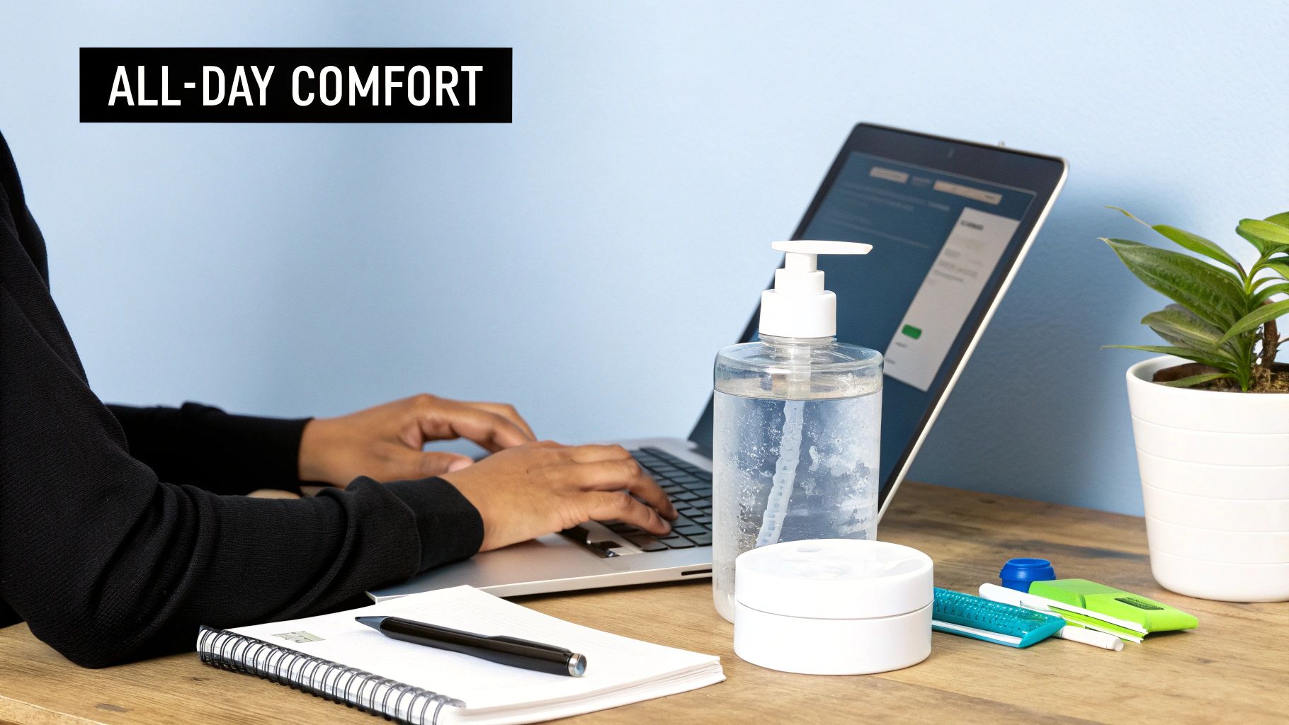 Person typing on a laptop at a neat workspace with hand sanitizer, a plant, and office supplies.