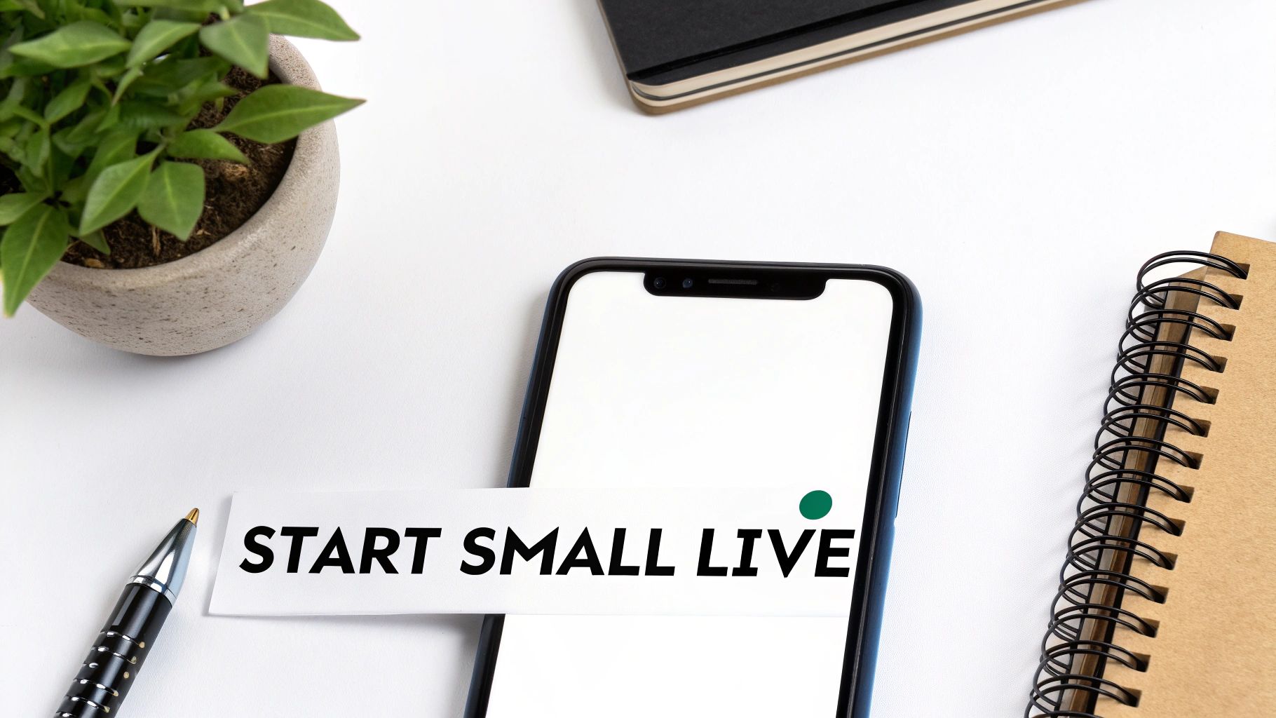 A flat lay of a white desk with a plant, pen, smartphone, and a 'START SMALL LIVE' note. A flat lay of a white desk with a plant, pen, smartphone, and a 'START SMALL LIVE' note.