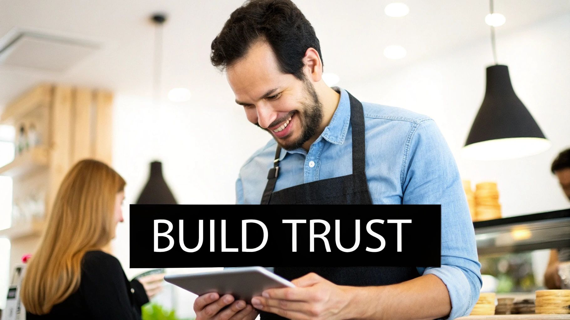 Smiling male barista in a cafe, looking at a tablet, with overlaid text 'BUILD TRUST'.