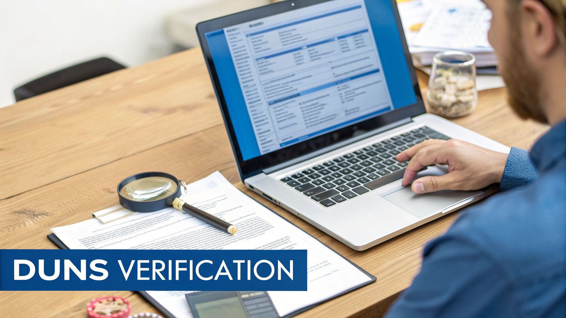 A person performing DUNS verification on a laptop, with documents and a magnifying glass on a wooden desk.
