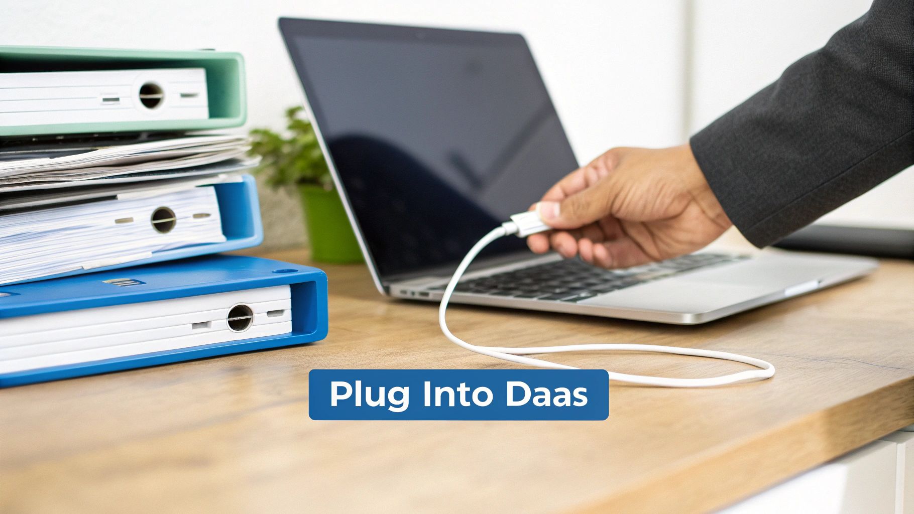 A hand plugs a white USB cable into a silver laptop on a wooden desk next to binders.