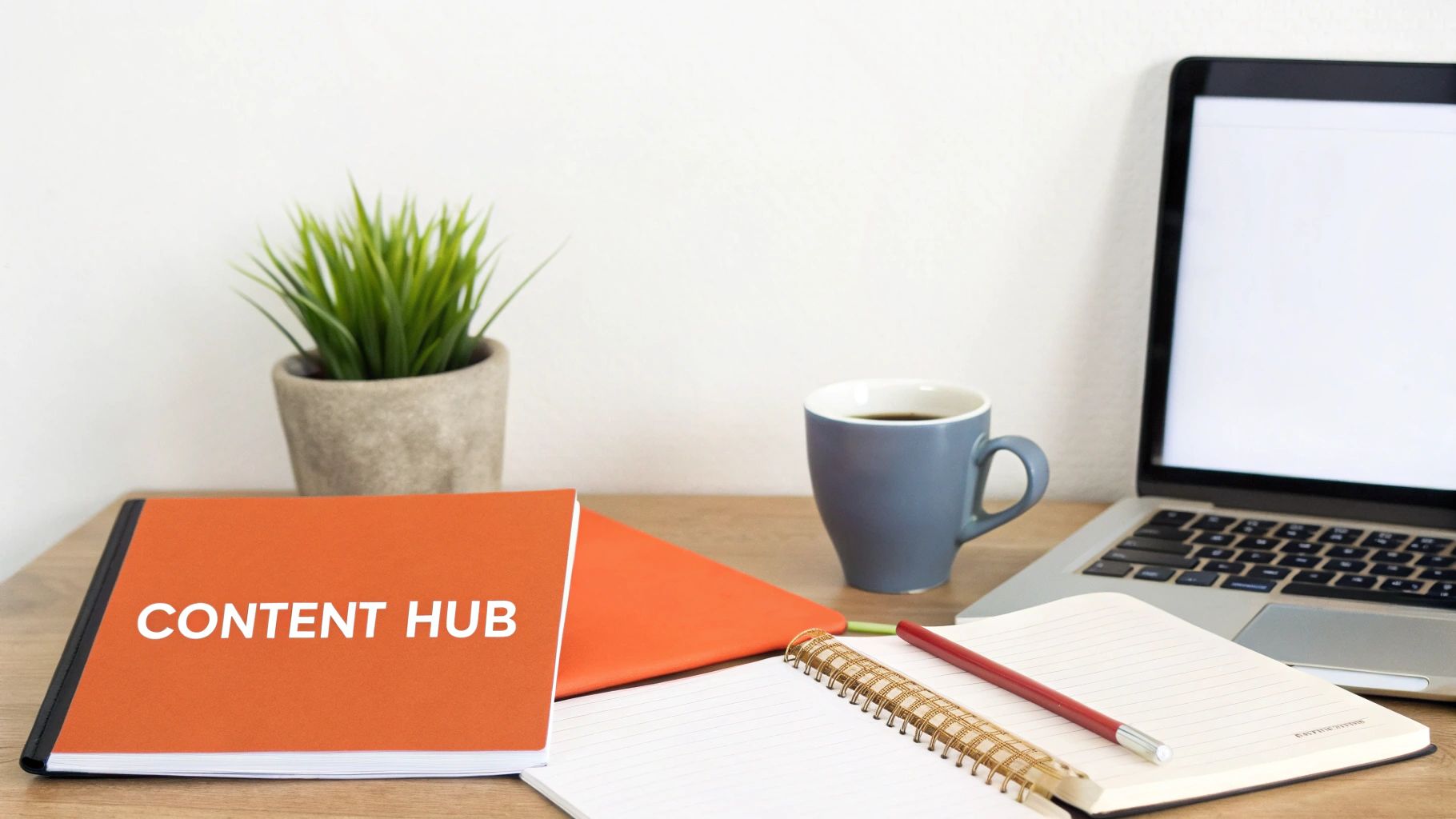 A clean workspace with an orange 'Content Hub' notebook, coffee, laptop, and plant on a wooden desk.