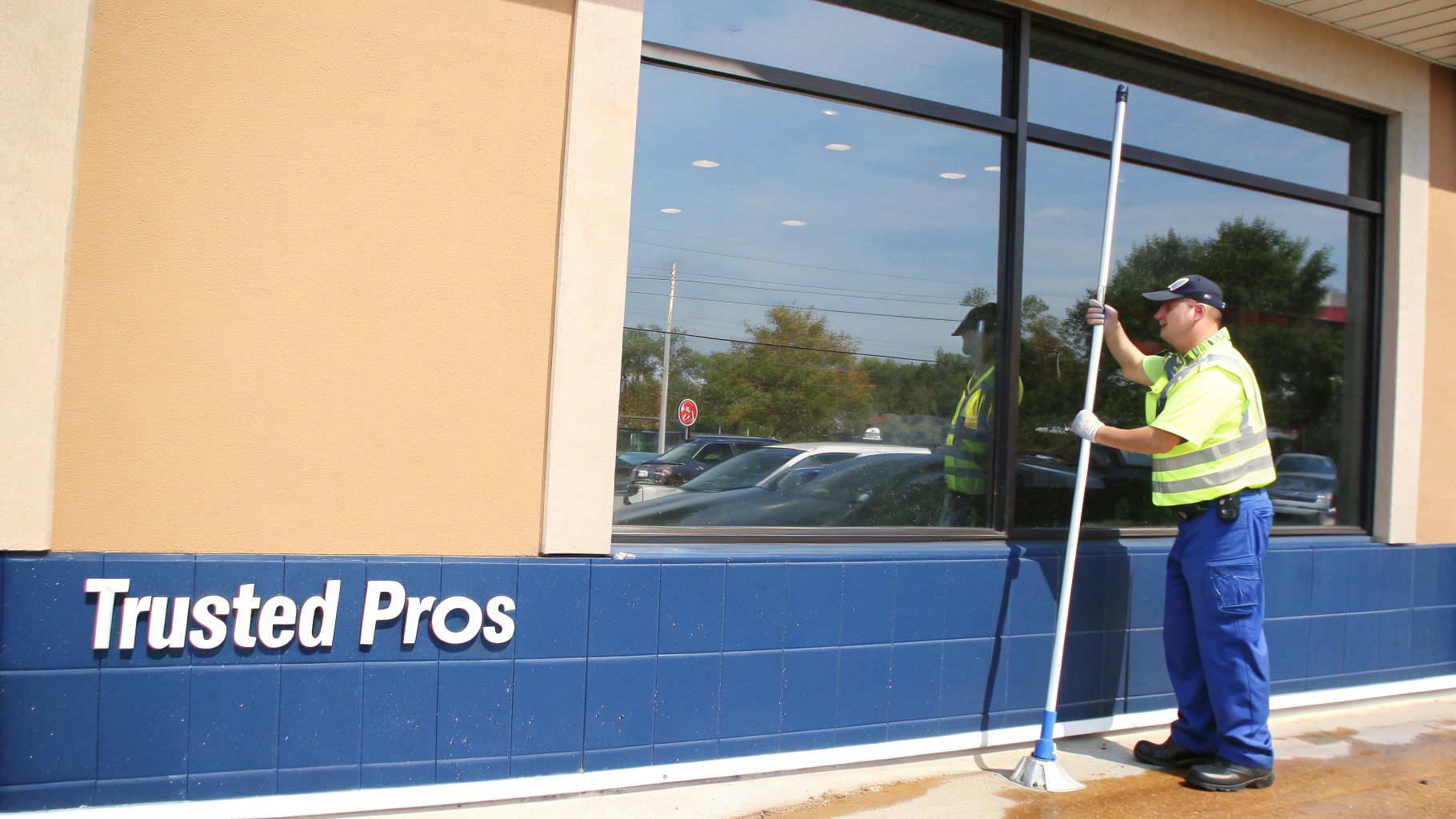 A professional window cleaner in a high-visibility vest uses a long brush to clean a large commercial window.