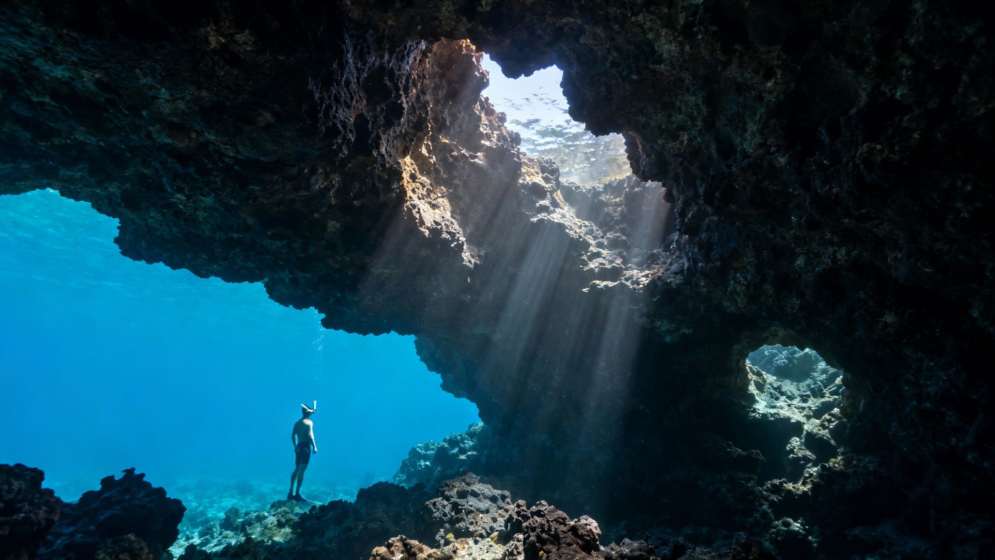A snorkeler stands in a vibrant blue underwater cave, illuminated by sunbeams from above.