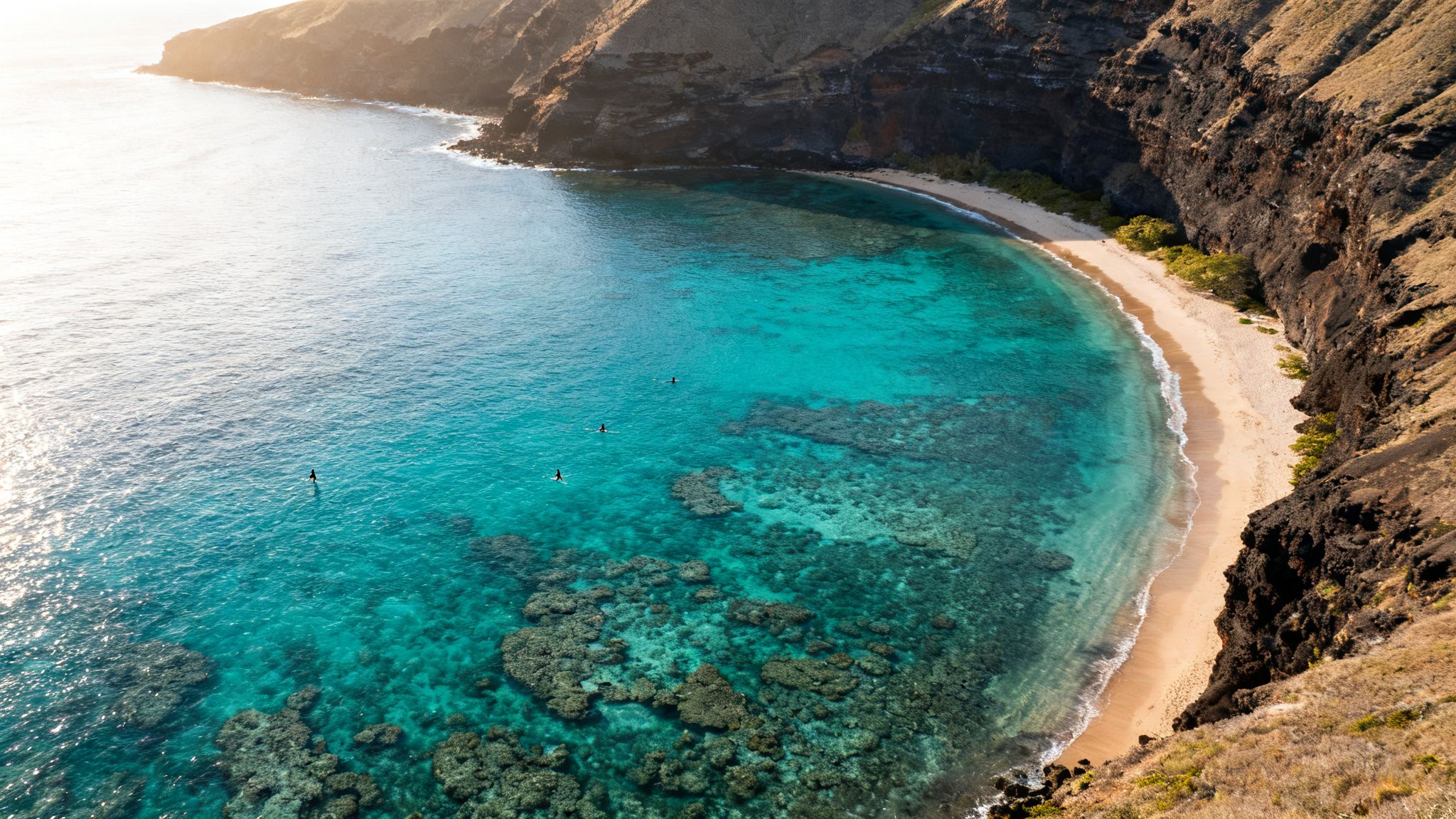 Aerial view of a secluded turquoise bay with coral reefs, a sandy beach, cliffs, and paddleboarders.