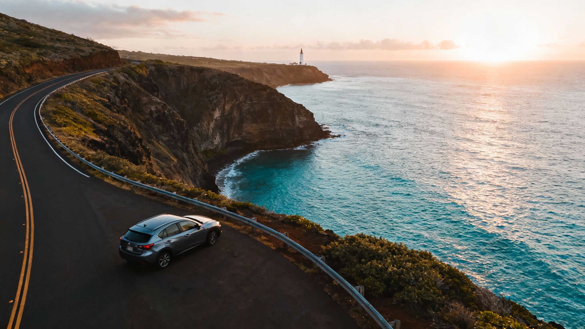 A car drives on a winding coastal road towards a lighthouse at sunset over the ocean.