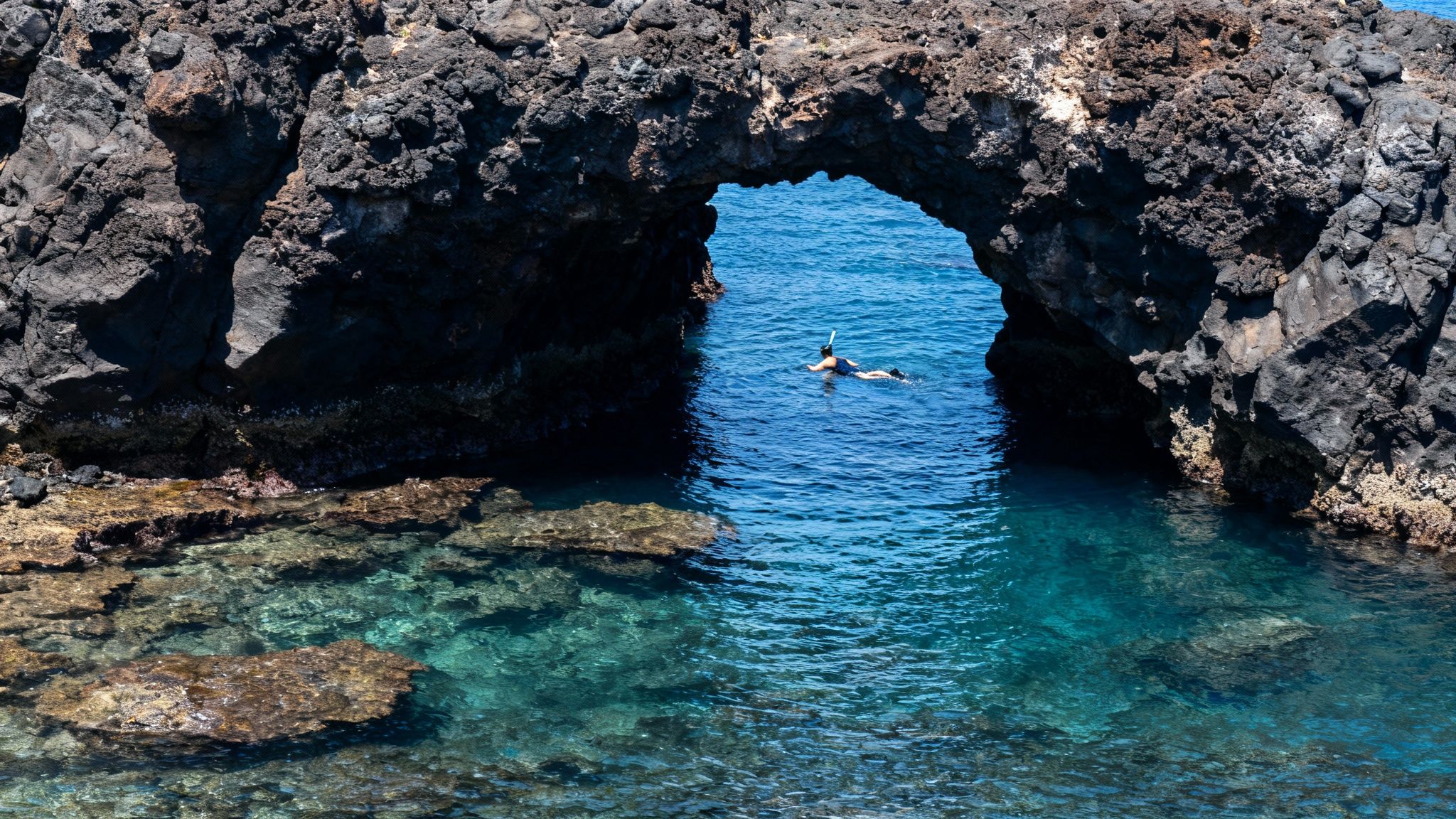 A person snorkeling through a natural rock arch in crystal-clear blue ocean water on a sunny day.