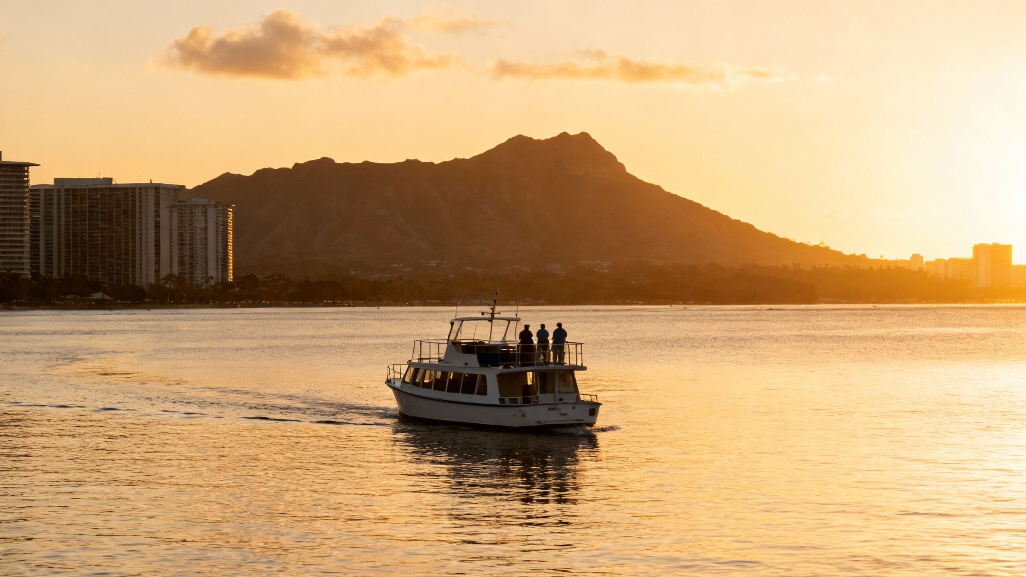 A boat carrying people sails on calm water during a golden sunset, with Diamond Head mountain and Honolulu buildings in the background.