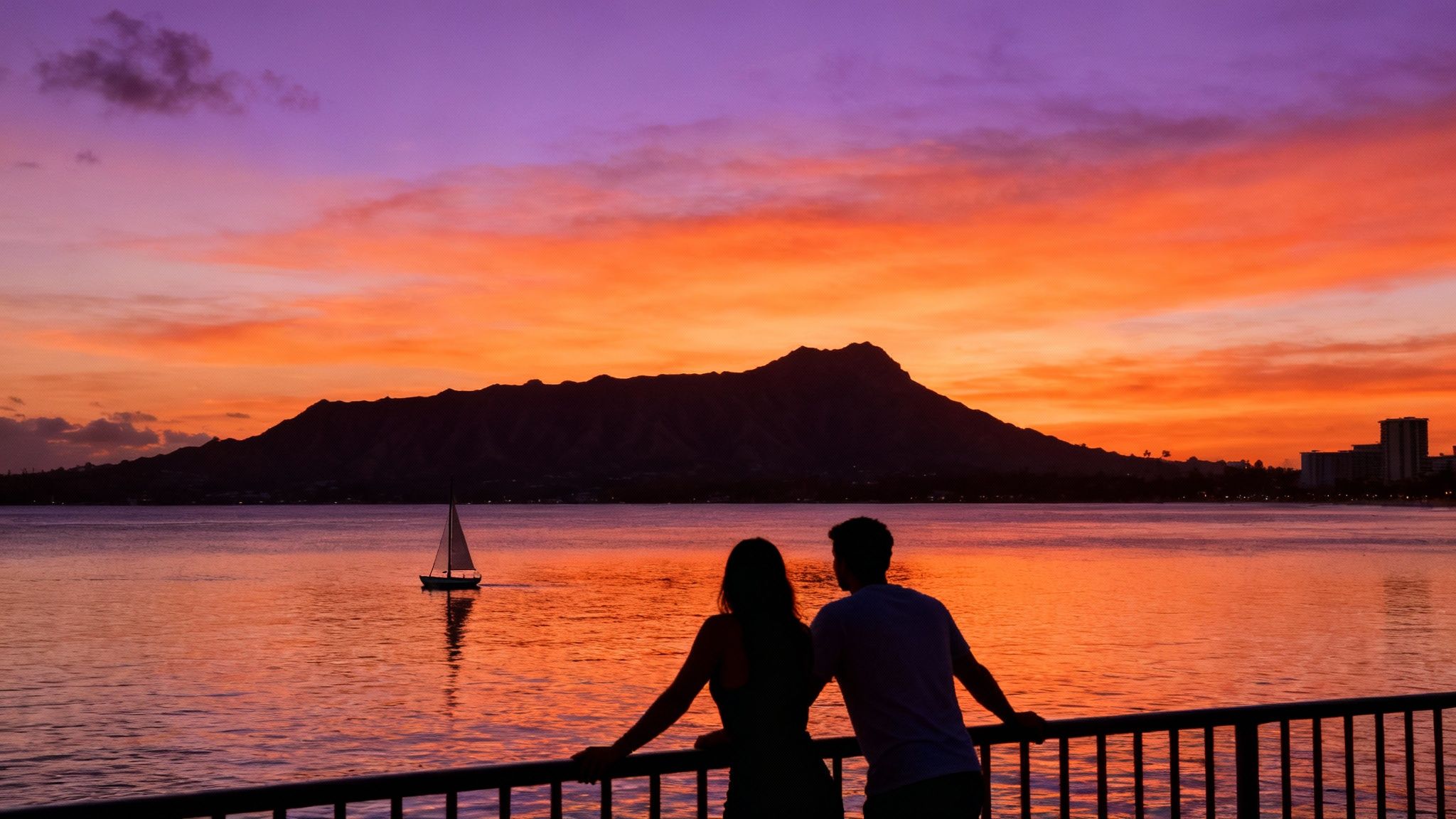 A couple watches a vibrant orange and purple sunset over the ocean with Diamond Head and a sailboat.