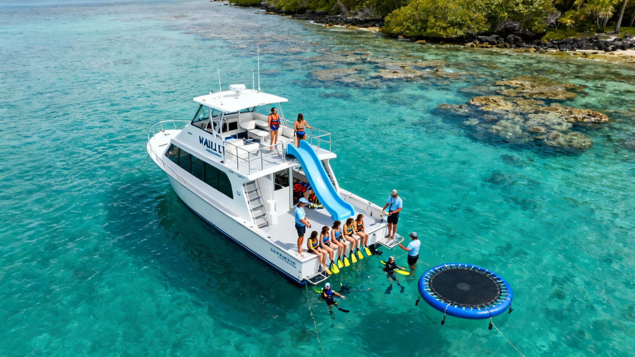 An aerial view of a white boat with a blue slide and people snorkeling in clear turquoise water near a lush coast.