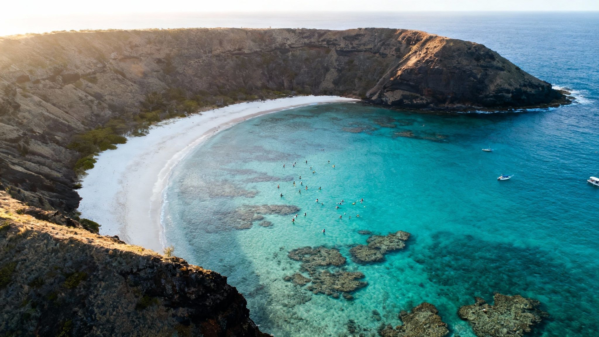 Aerial view of a stunning turquoise bay with a white sand beach, cliffs, many swimmers, and boats.