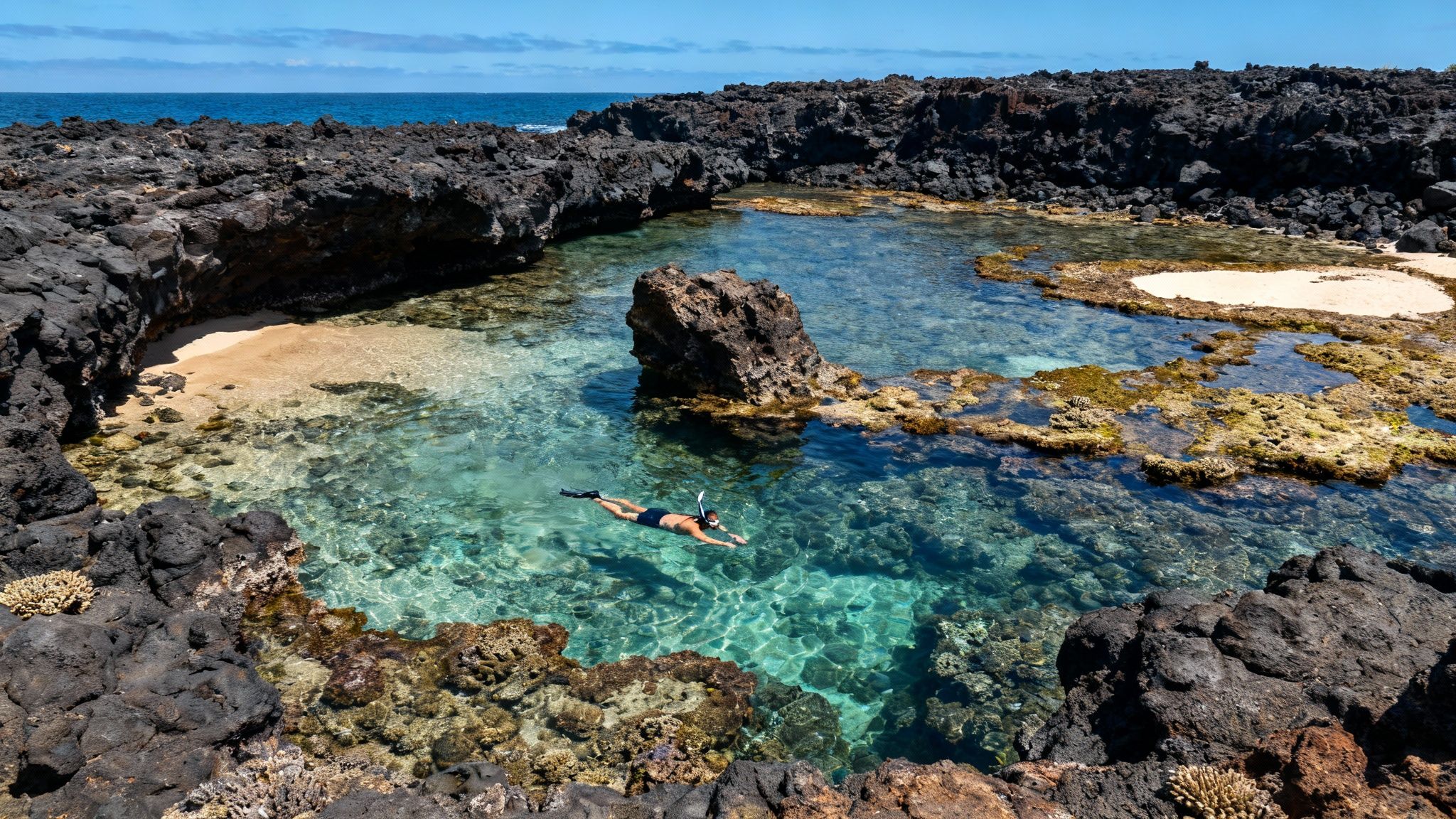 Person snorkeling in a clear blue lagoon surrounded by dark volcanic rocks and sandy beach.