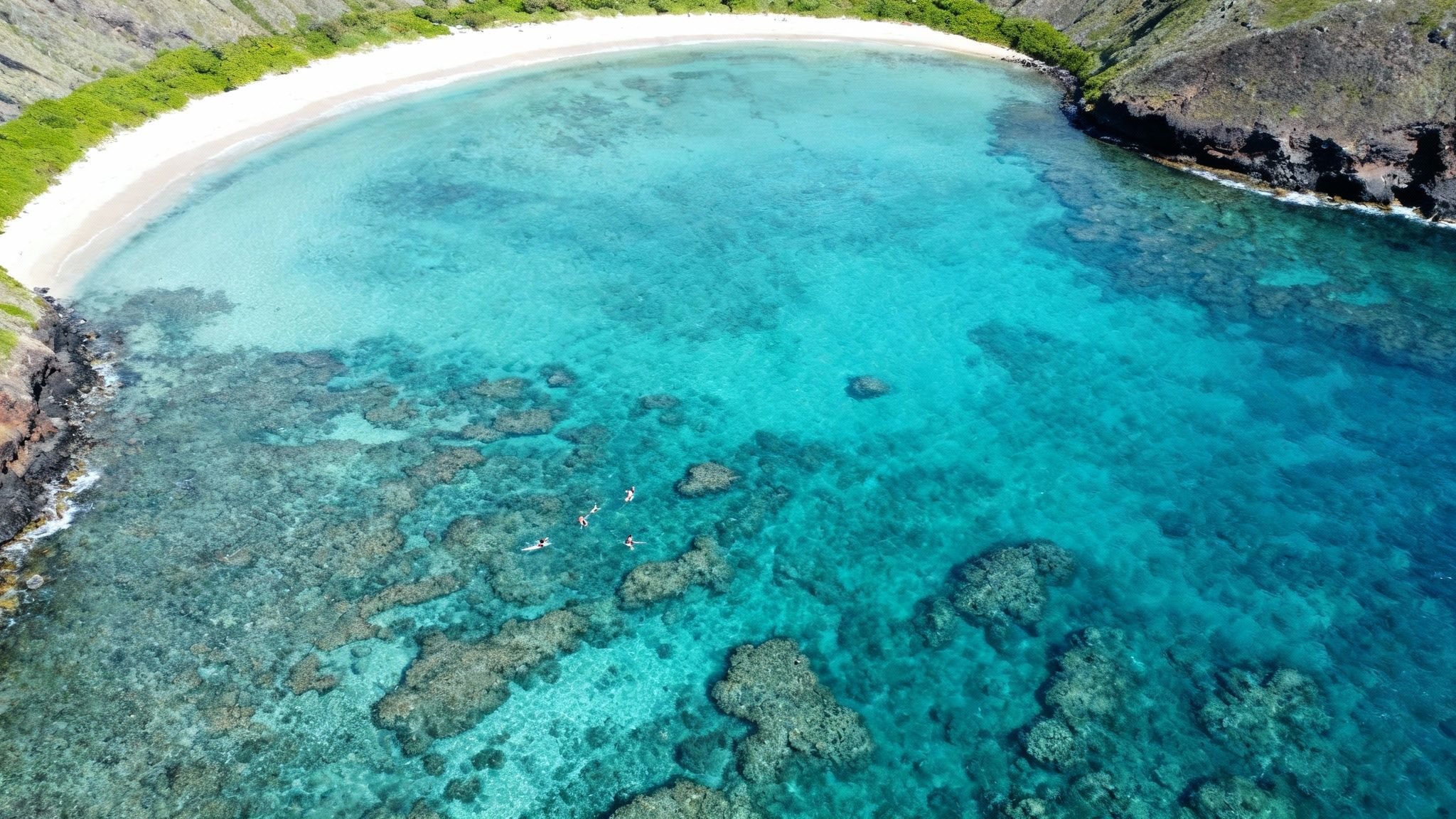 Aerial view of a stunning tropical bay with clear turquoise water, white beach, and people snorkeling over vibrant coral.
