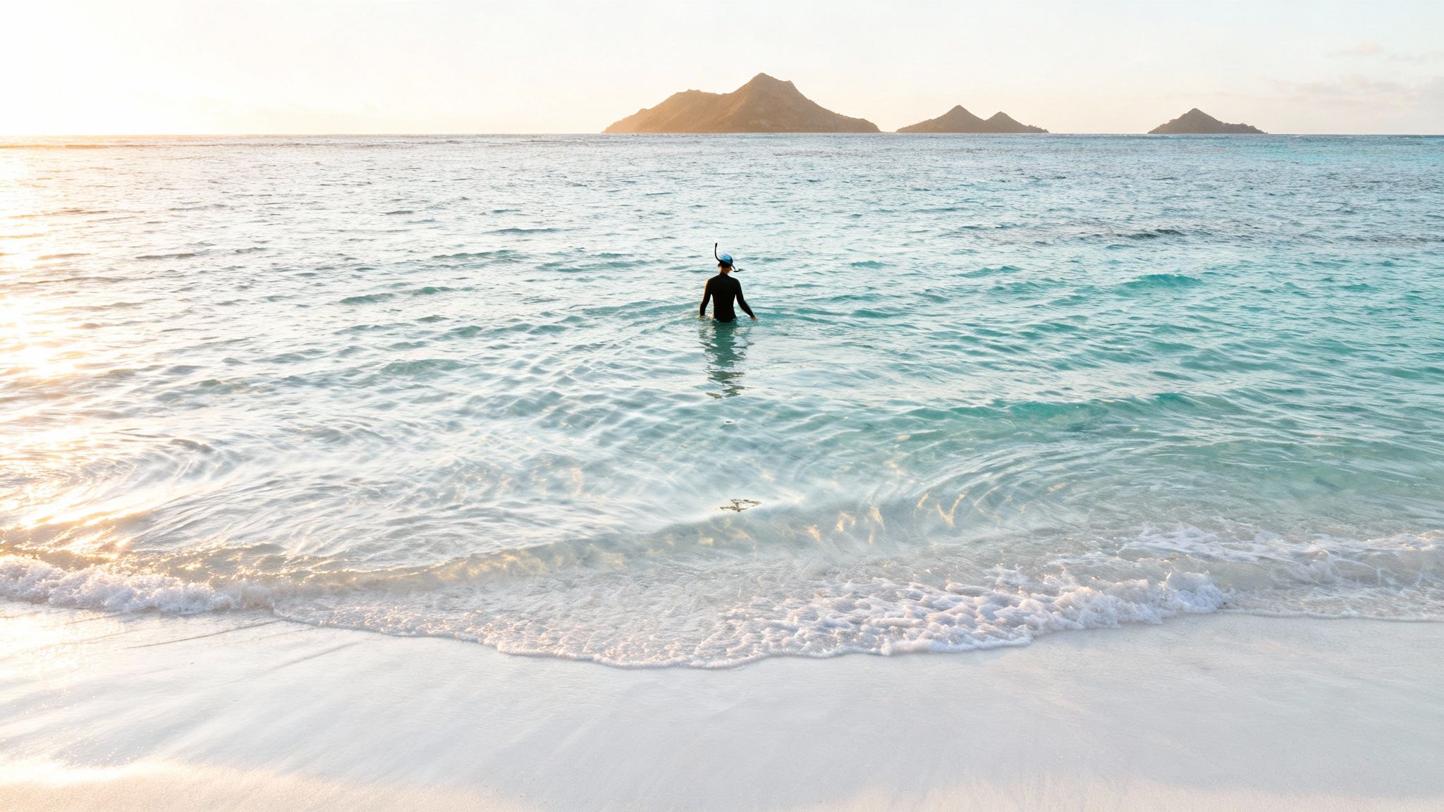 A person in a wetsuit and snorkel stands in calm, clear turquoise ocean water at sunset, facing distant islands.