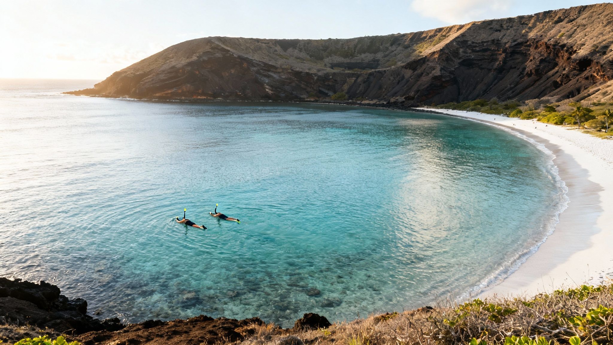 Two people snorkeling in clear turquoise water near a white sand beach and volcanic crater.