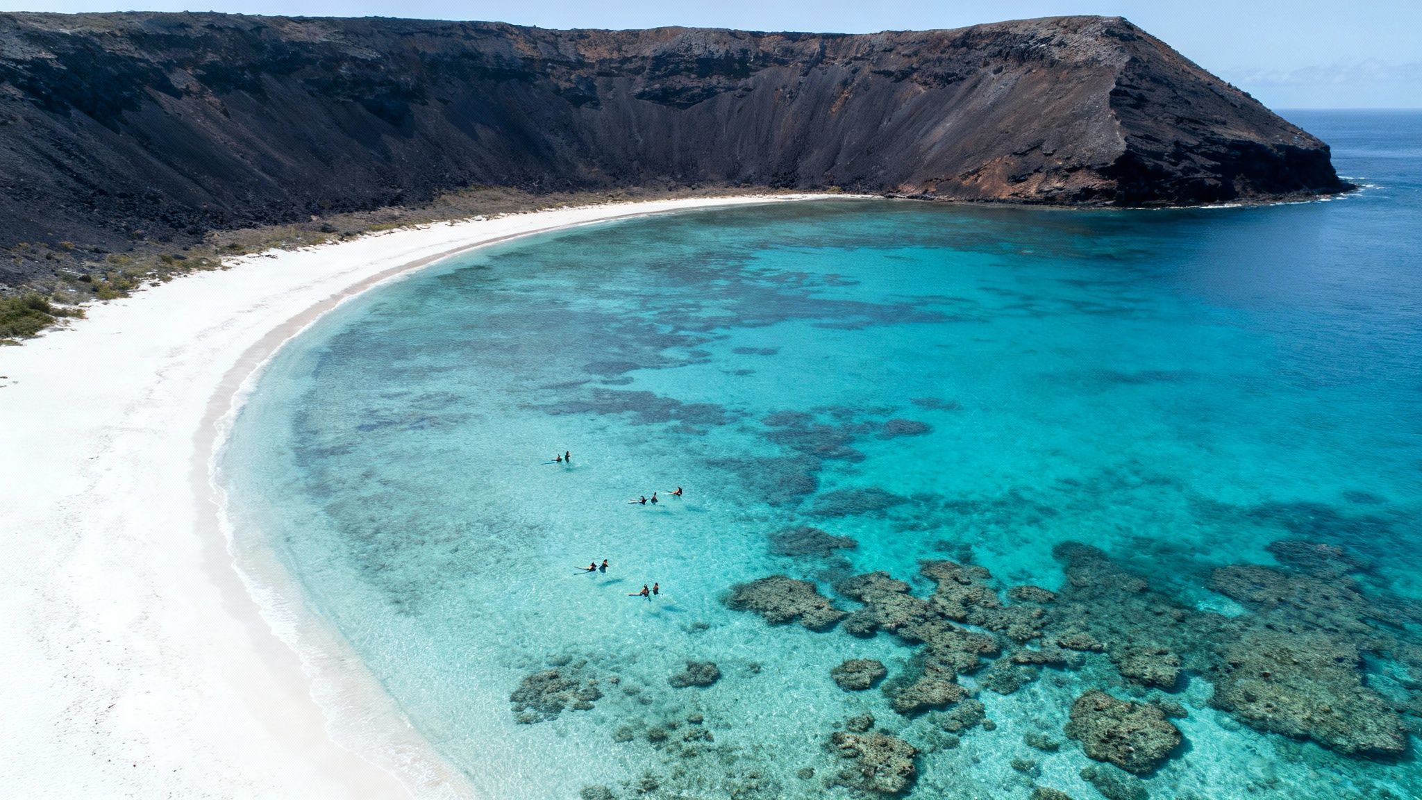 Aerial view of a stunning tropical beach with white sand, clear turquoise water, dark cliffs, and people snorkeling.