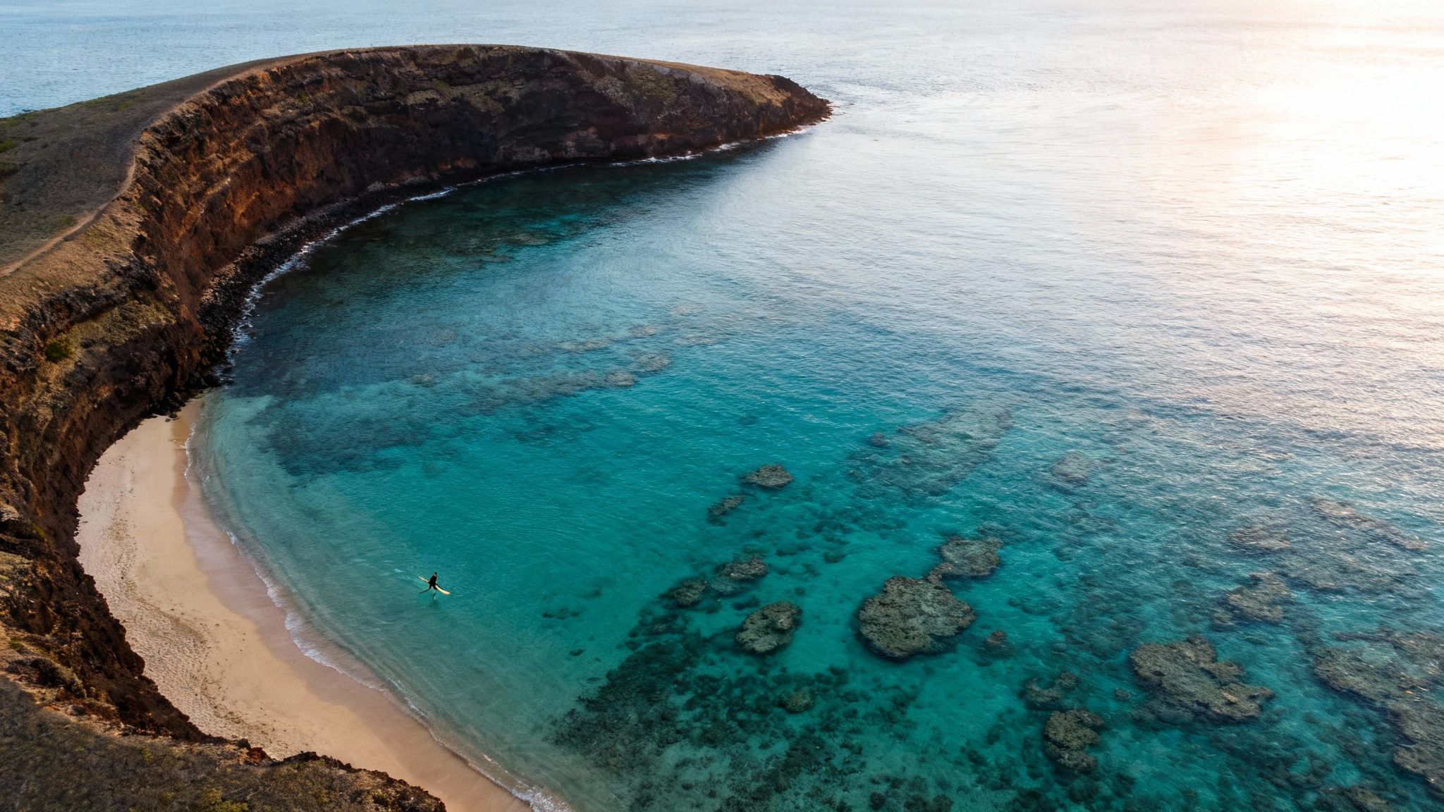 Aerial view of a person on a paddleboard in clear turquoise water beside a sandy beach and cliffs.