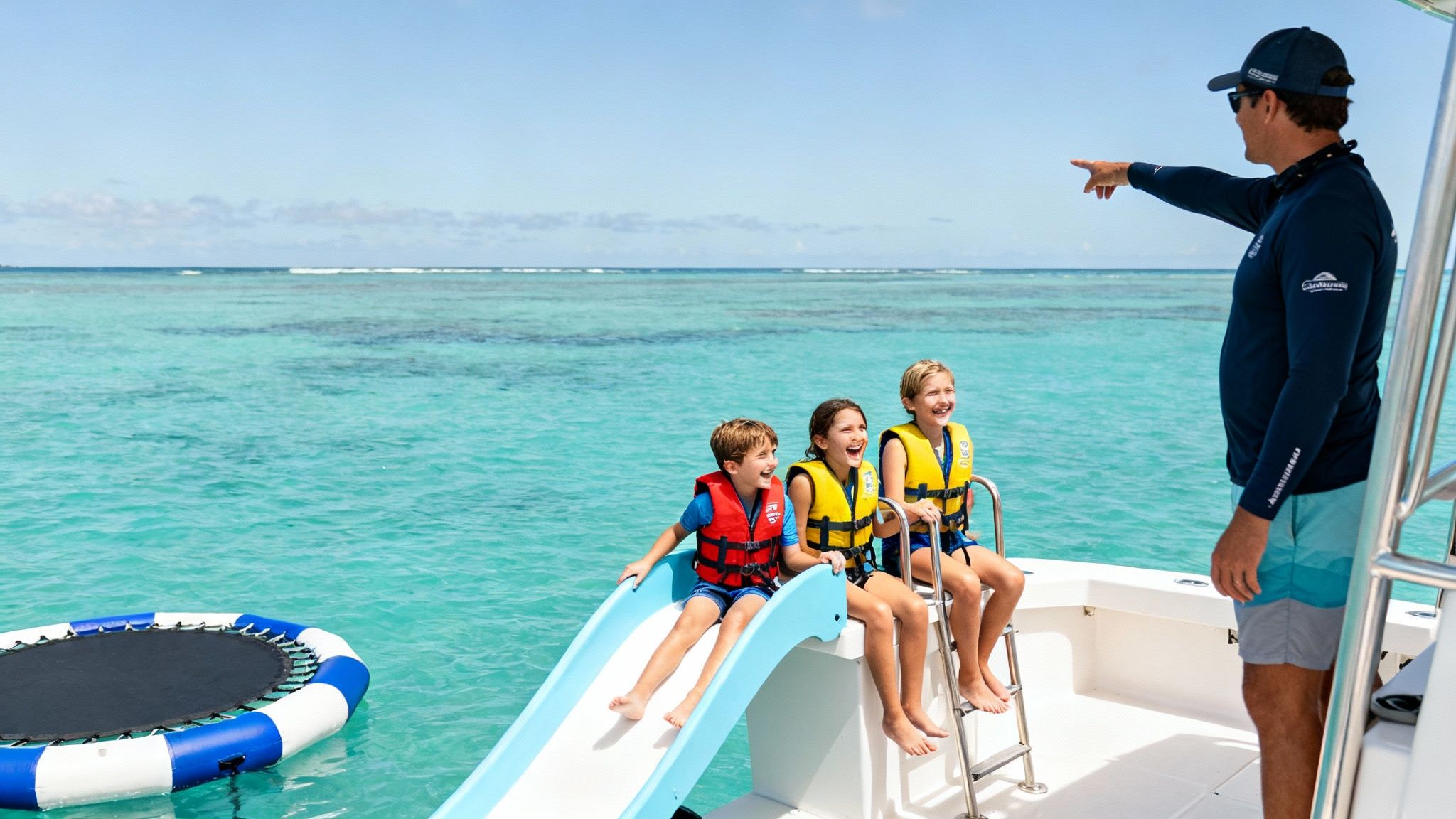 A man points to the ocean while three smiling children in life vests enjoy a boat day with a slide.