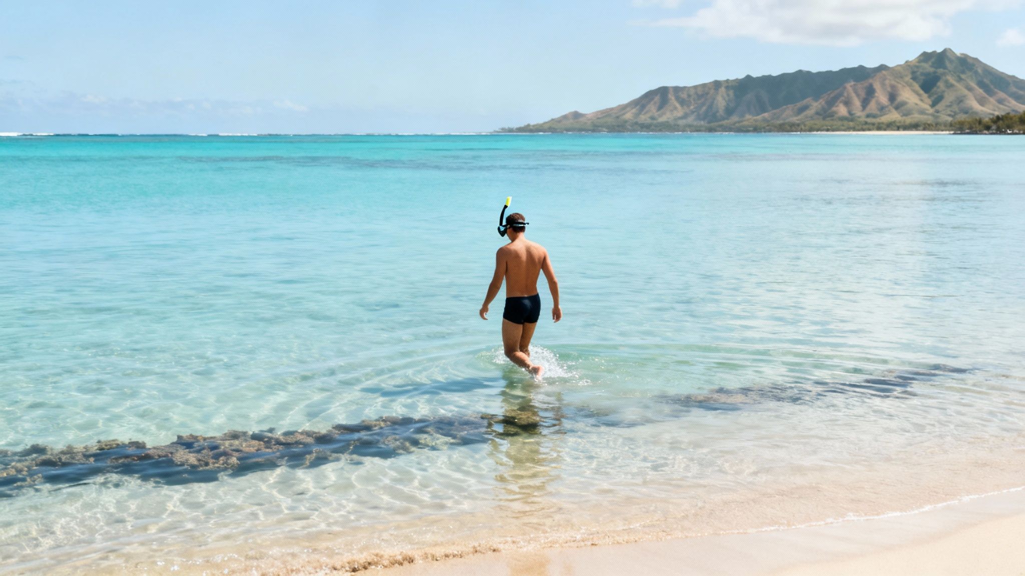 A man in black swim trunks with a snorkel and mask walks into clear blue ocean water from a sandy beach, with mountains in the background.