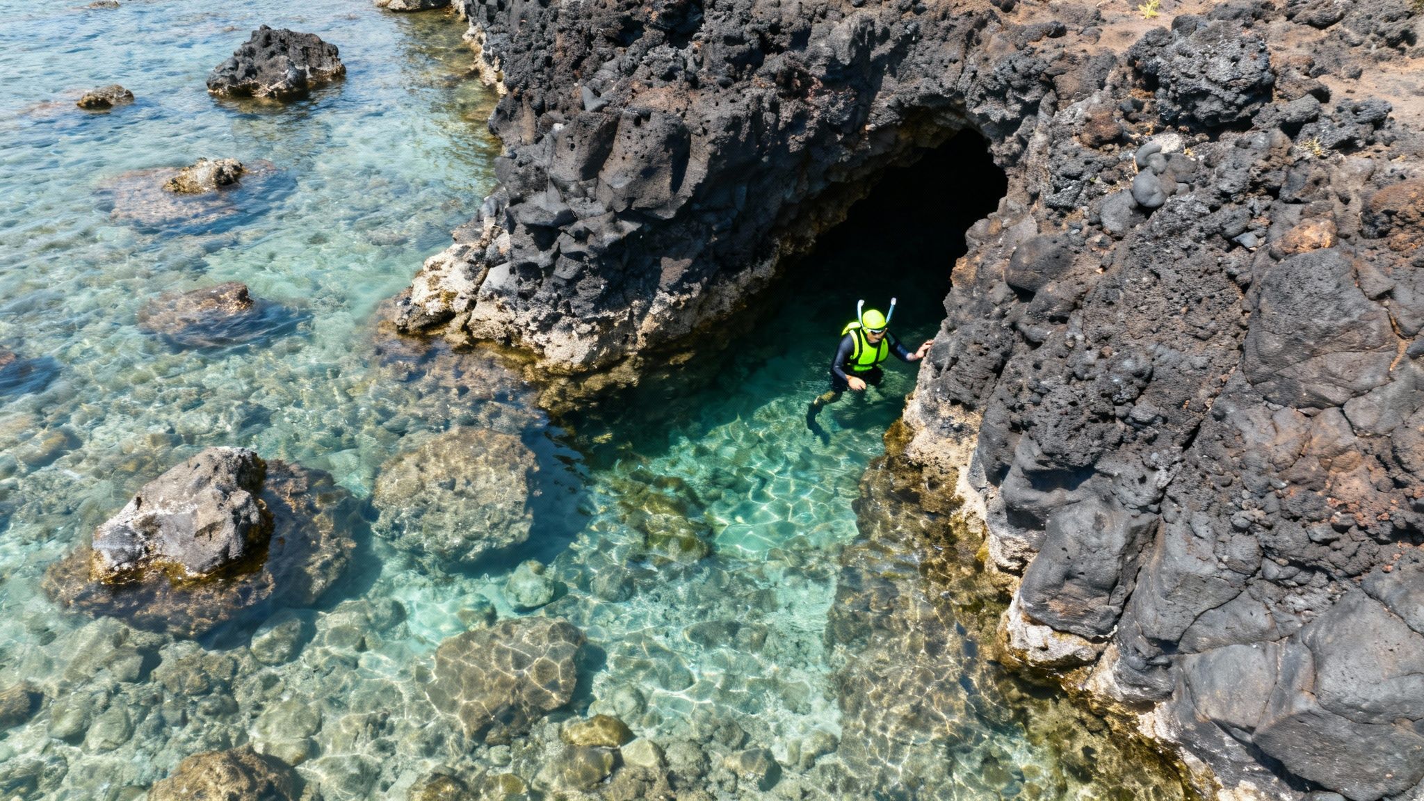 Person in snorkeling gear exploring a clear turquoise water cave entrance with dark volcanic rocks.