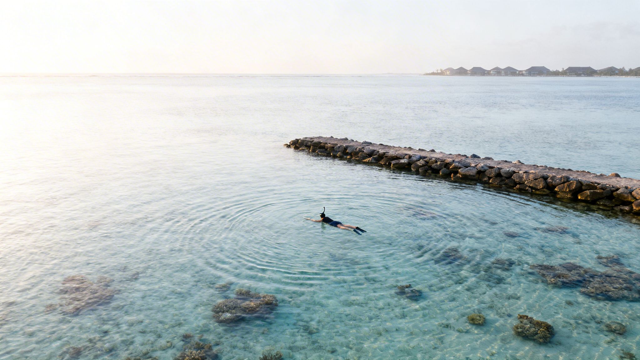 A person snorkeling in clear blue water near a rocky pier with bungalows in the distance.